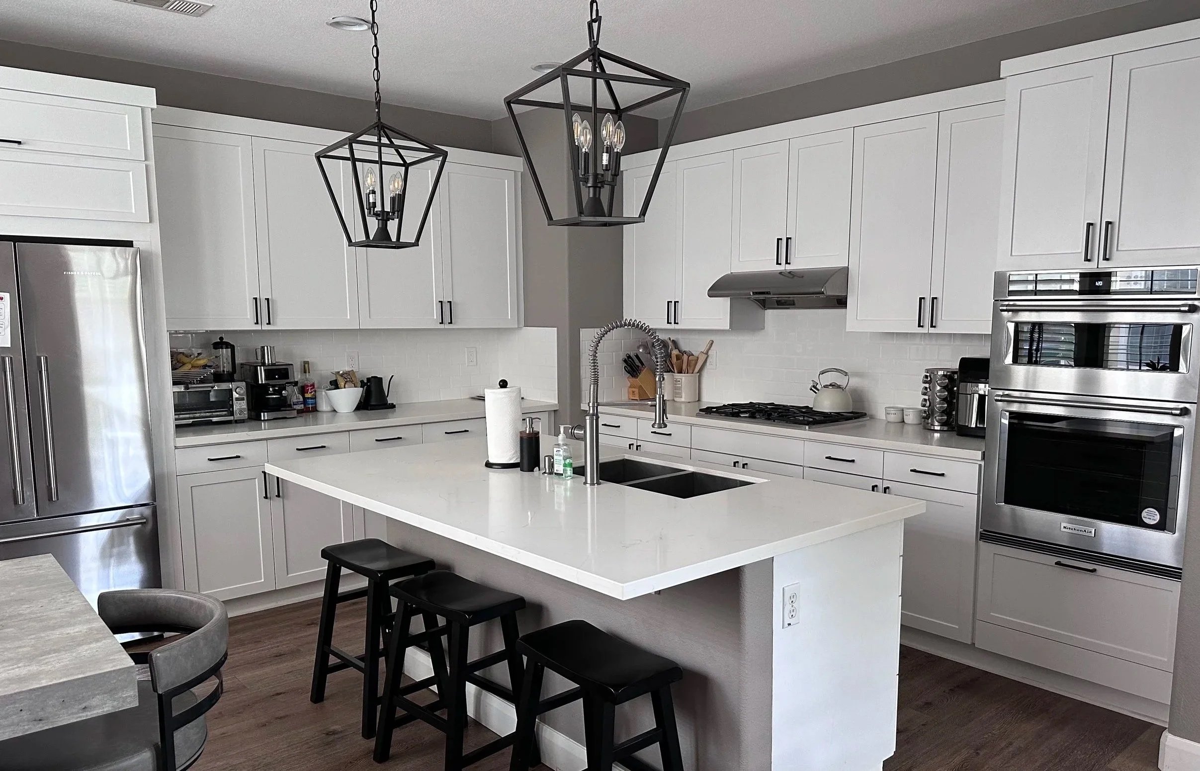 A modern kitchen with white cabinets, a white countertop island, stainless steel appliances including a refrigerator and double oven, and two black geometric pendant light fixtures hanging from the ceiling.