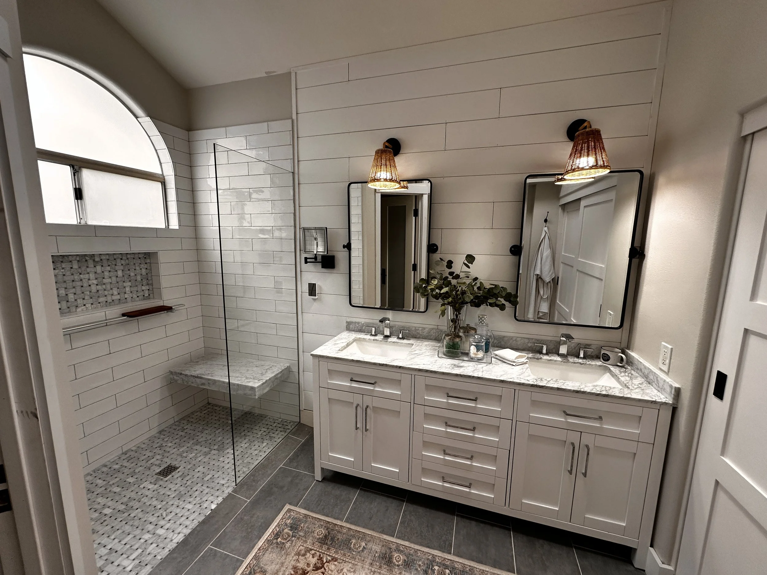 Modern bathroom with white shiplap walls, dual mirrors, two lamps with woven shades, a marble countertop with sinks, a glass shower with white subway tiles, a small plant, and various toiletries.