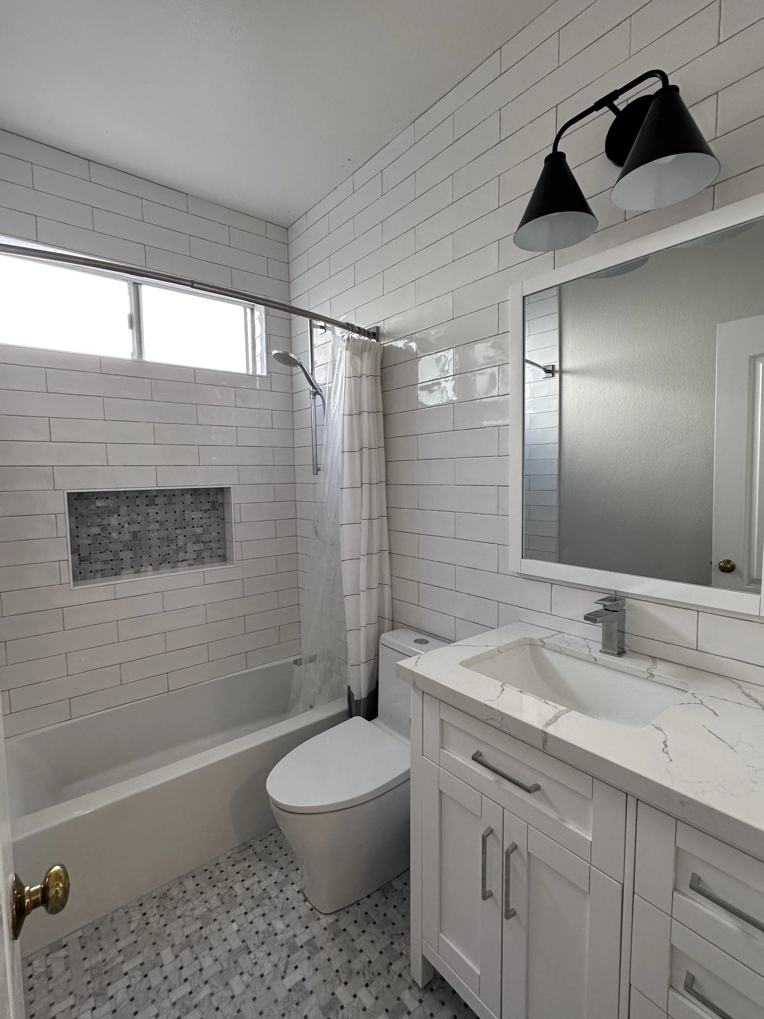 Clean modern bathroom with white tile walls, a bathtub with a showerhead and a curtain, a white toilet, a white vanity with a marble countertop and a rectangular mirror, and a black wall-mounted light fixture.