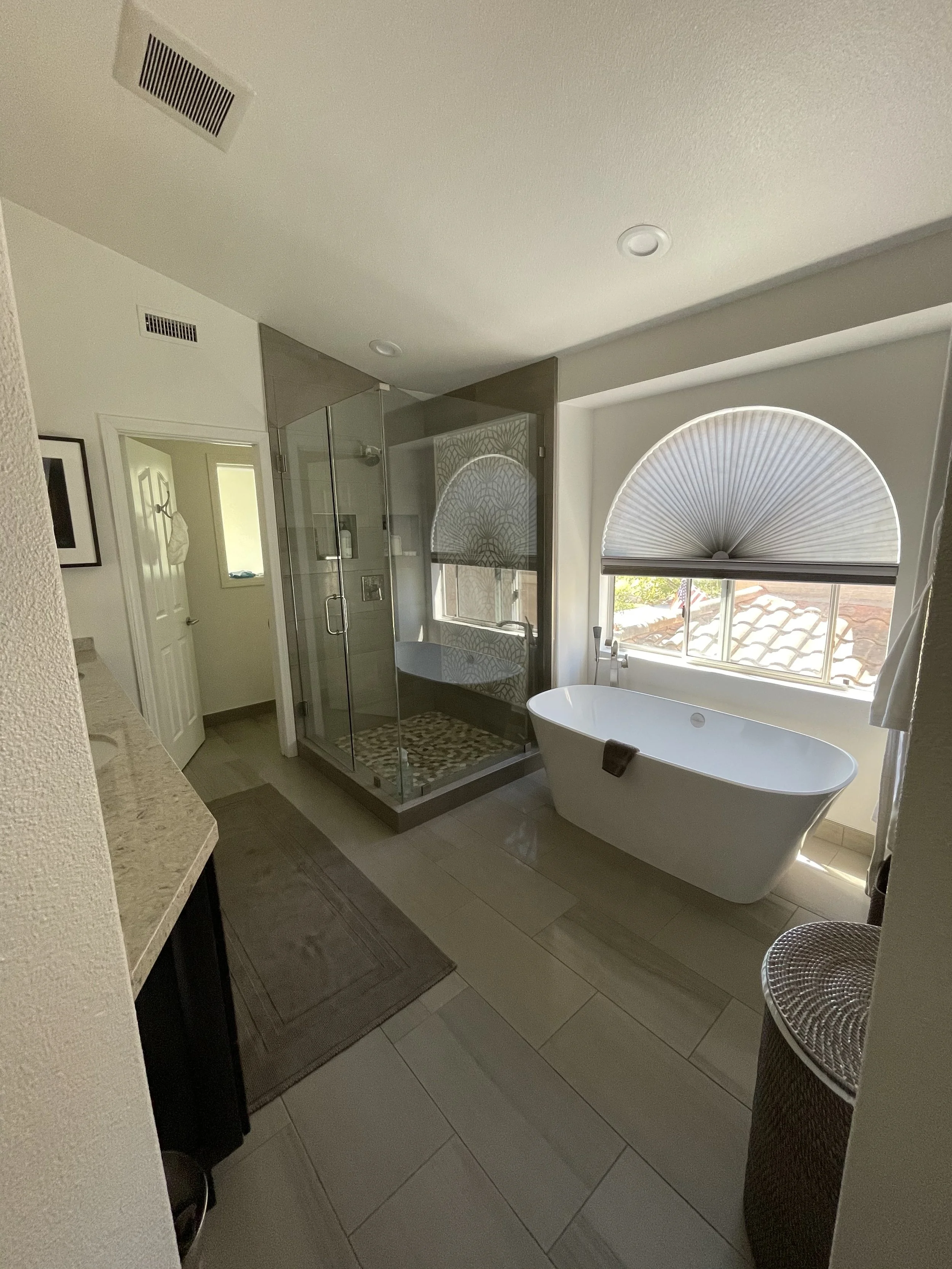 Bathroom with a freestanding bathtub, glass shower enclosure, window with half-circle shade, wicker chair, and tiled floor.