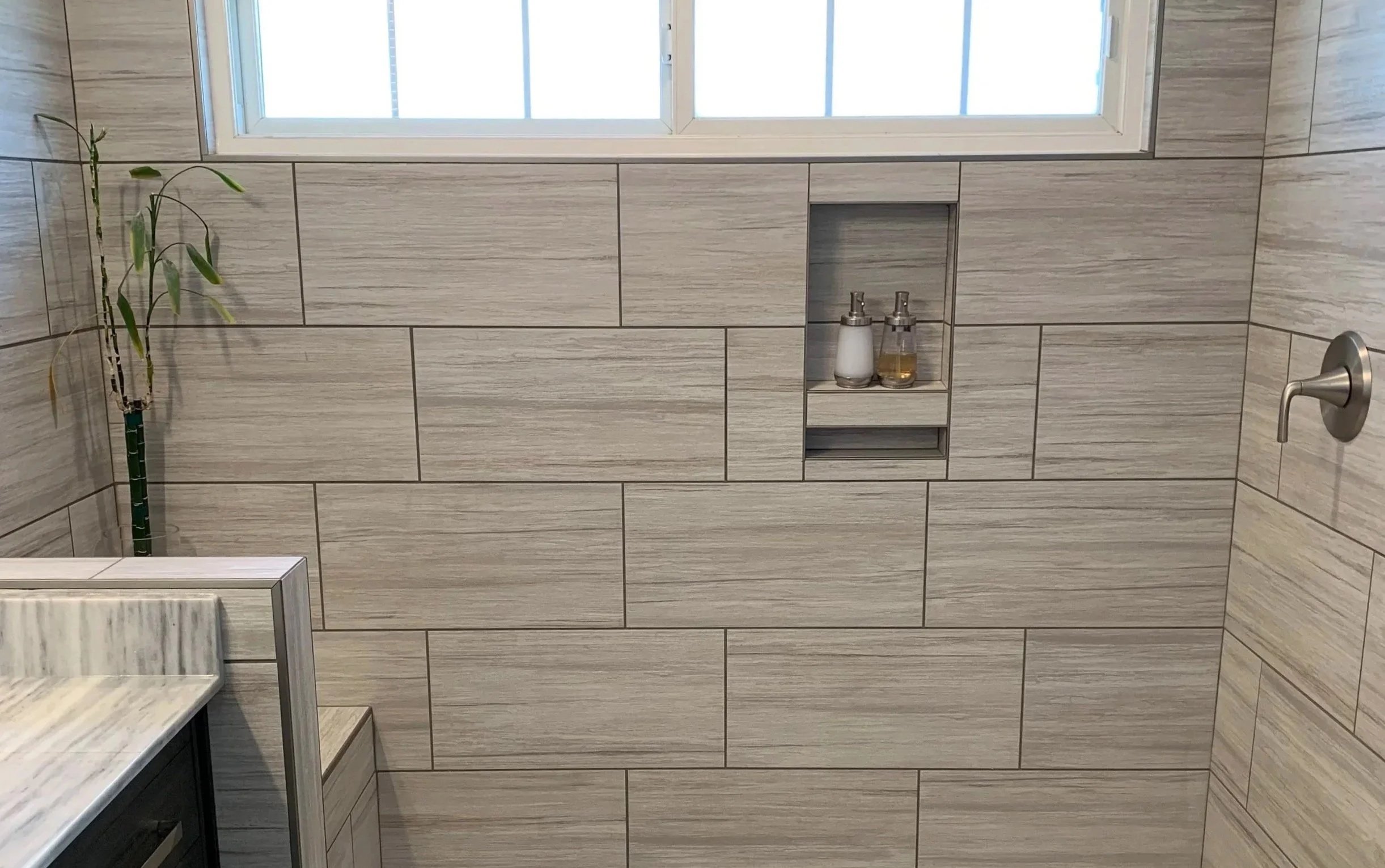 A bathroom with beige horizontal subway tiles, a small built-in shelf with soap dispensers, a window at the top, a potted plant to the left, and a marble countertop edge in the bottom left corner.