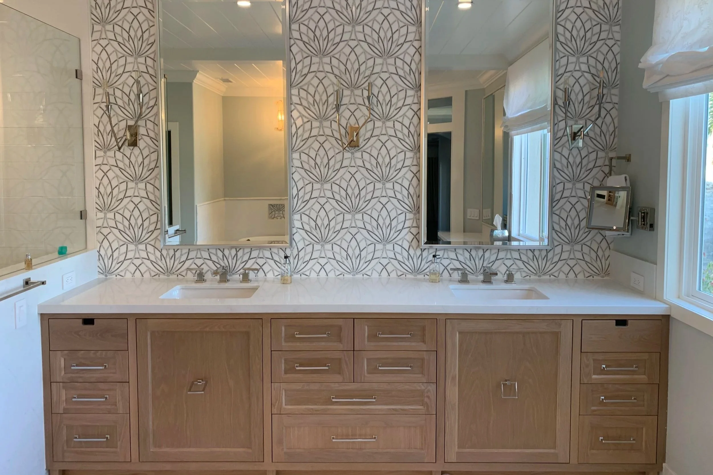 A modern bathroom vanity area with a large white countertop, two sinks, and eight wooden drawers. Behind the countertop, there are two large vertical mirrors with decorative wall panels featuring a geometric leaf pattern. To the right, there are two windows with white blinds, and to the left, outside the mirror, there is a glimpse of a light wall with a wall sconce.