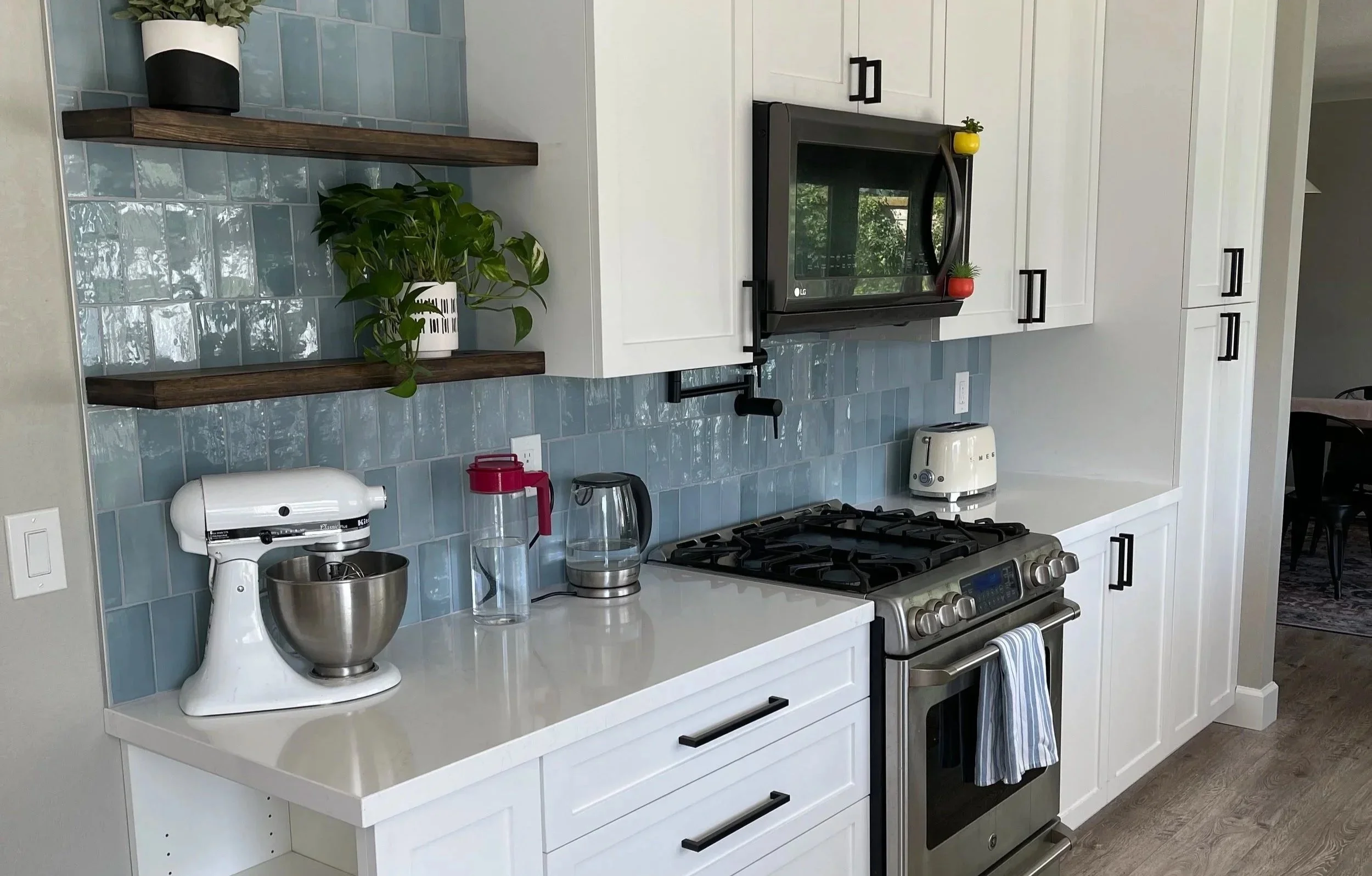 Kitchen with white cabinets, a gray stove and oven, a black microwave, wooden shelves with potted plants, a blue tile backsplash, and various appliances on the countertop including a mixer, a toaster, and water bottles.