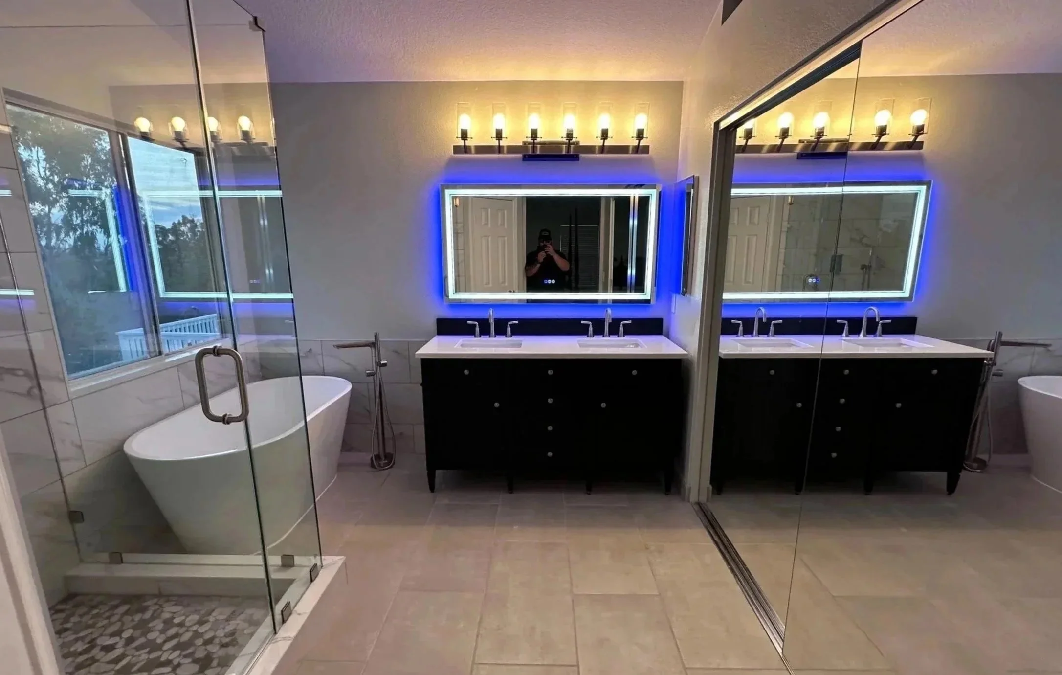 Modern bathroom with double vanity mirrors surrounded by blue LED lighting, a bathtub in the corner, and a walk-in shower with glass doors.