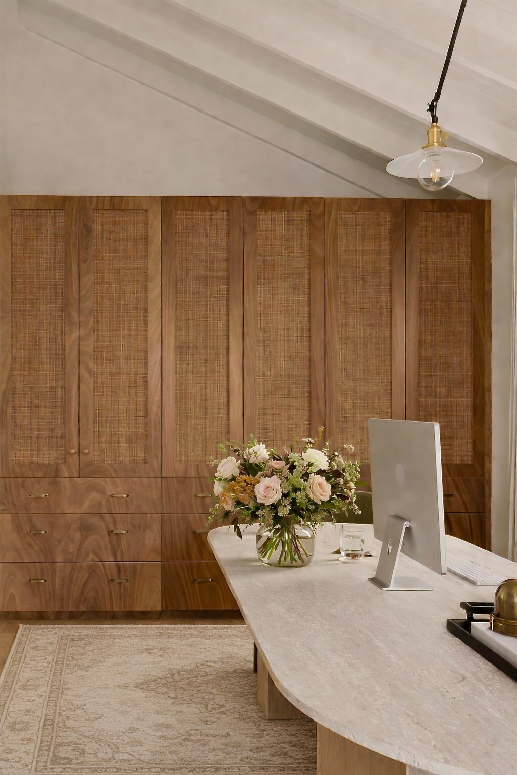 Interior of an office with a wooden cabinet, a white marble table, a computer monitor, a glass of water, a bouquet of flowers, and a pendant light fixture.