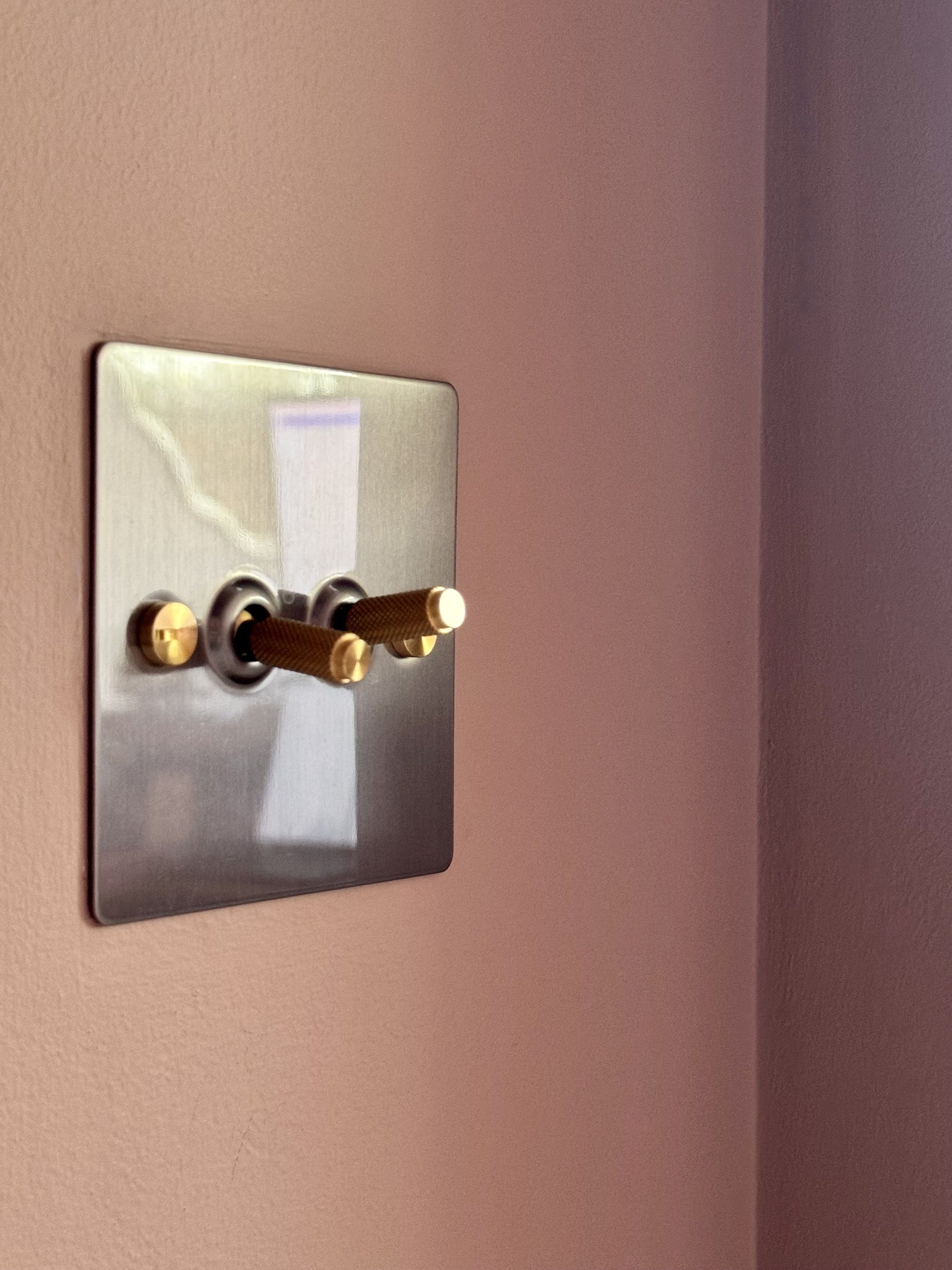 Close-up of a metal electrical switch plate with three switches on a pink wall.