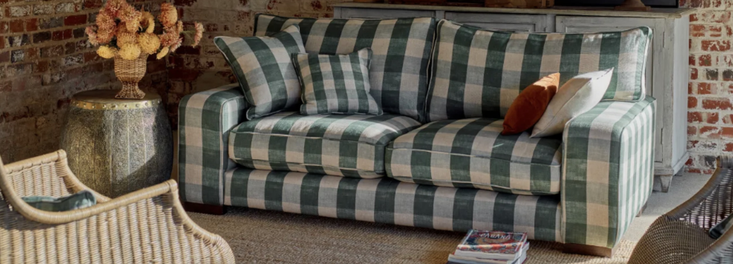 Living room with a green and beige checkered sofa, a wicker chair, a floral arrangement on a side table, and a brick wall background.