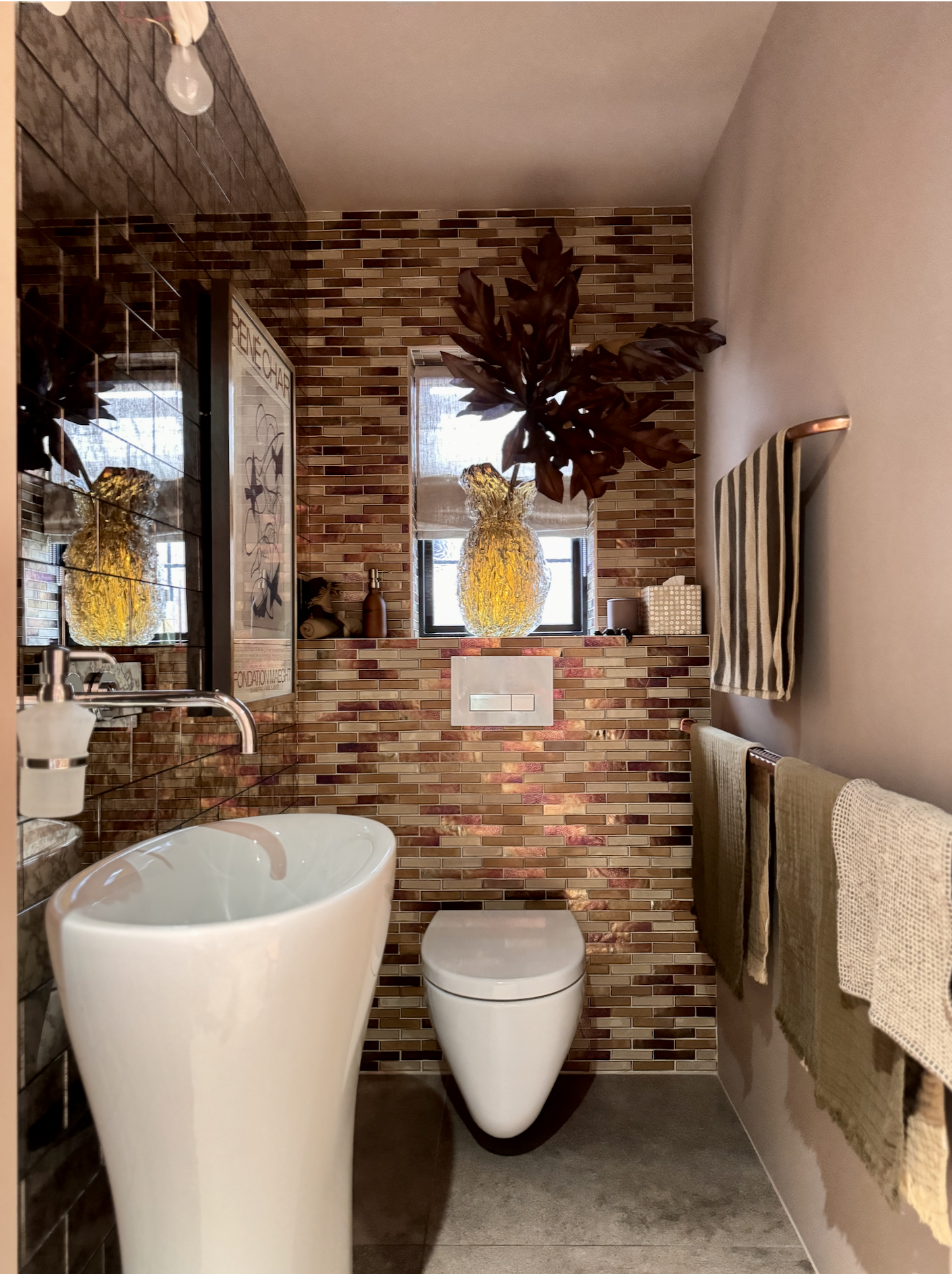 A modern guest cloakroom with a brick patterned wall tiles, a tall white sink, a wall-mounted toilet, a window with a large decorative vase on the windowsill, and towels hanging on hooks.