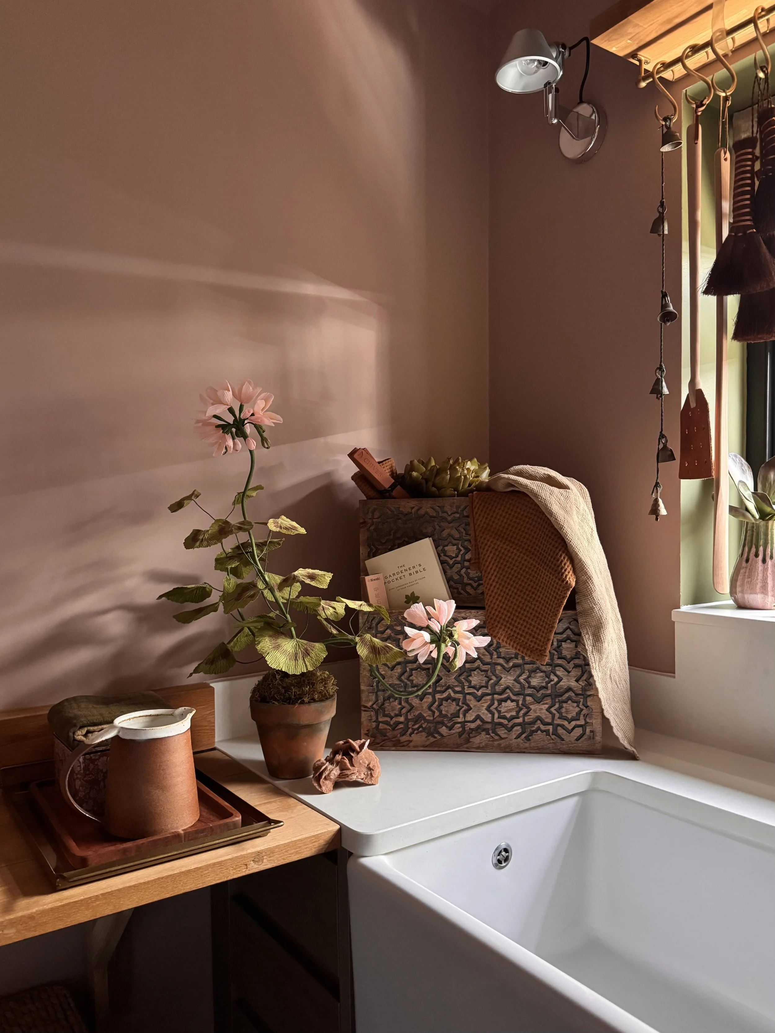 Interior of a kitchen with a white sink, wooden countertop, potted pink flowers, decorative jars, and a basket with green artichokes. There is a wall with shelves holding hanging wooden utensils and a small window with sunlight streaming in.