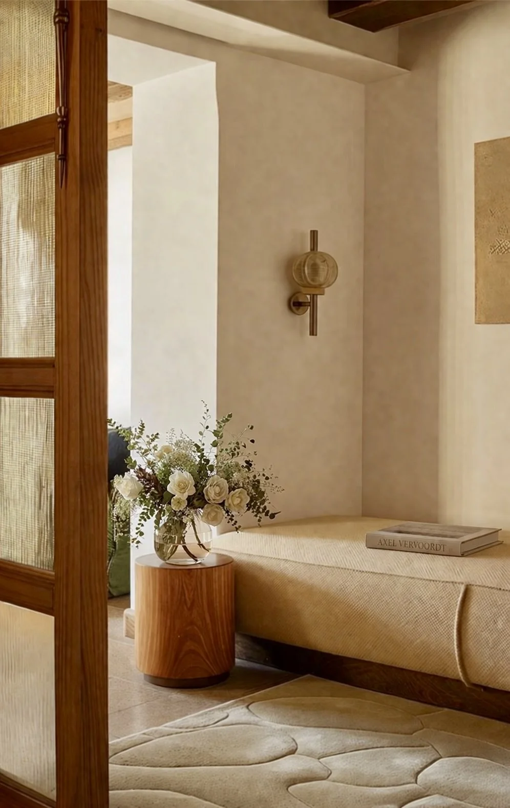 A cozy corner of a room featuring a beige daybed with a book on top, a wooden side table with a vase of white roses and greenery, a wall-mounted brass lamp, and a textured cream-colored wall with an abstract art piece. A wooden-framed door and a patterned area rug are also visible.