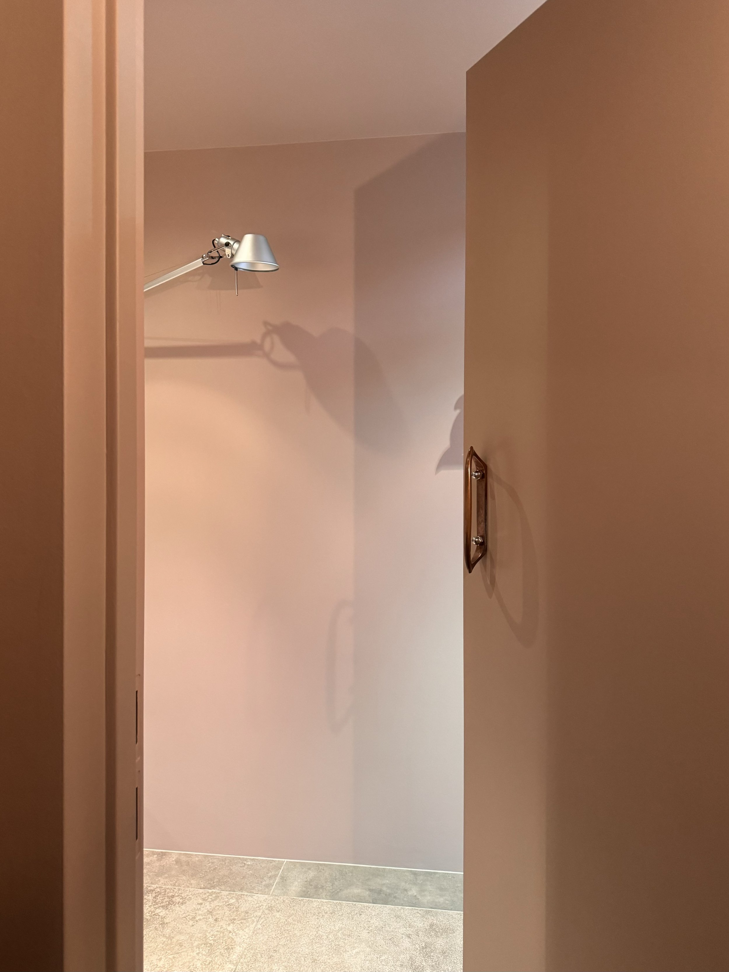 View of an indoor hallway seen through an open door, featuring beige walls, a metal wall-mounted lamp, and tiled floor.