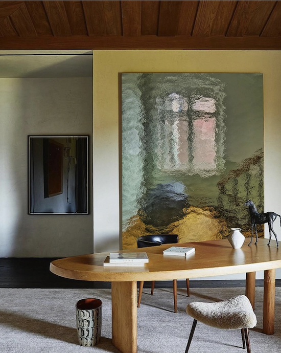 A dining room with an oval wooden table, a beige upholstered chair, and decorative items including a white vase and a horse figurine. A large abstract painting with a window scene hangs on the wall behind the table. Nearby, a flat-screen TV is mounted on the wall.