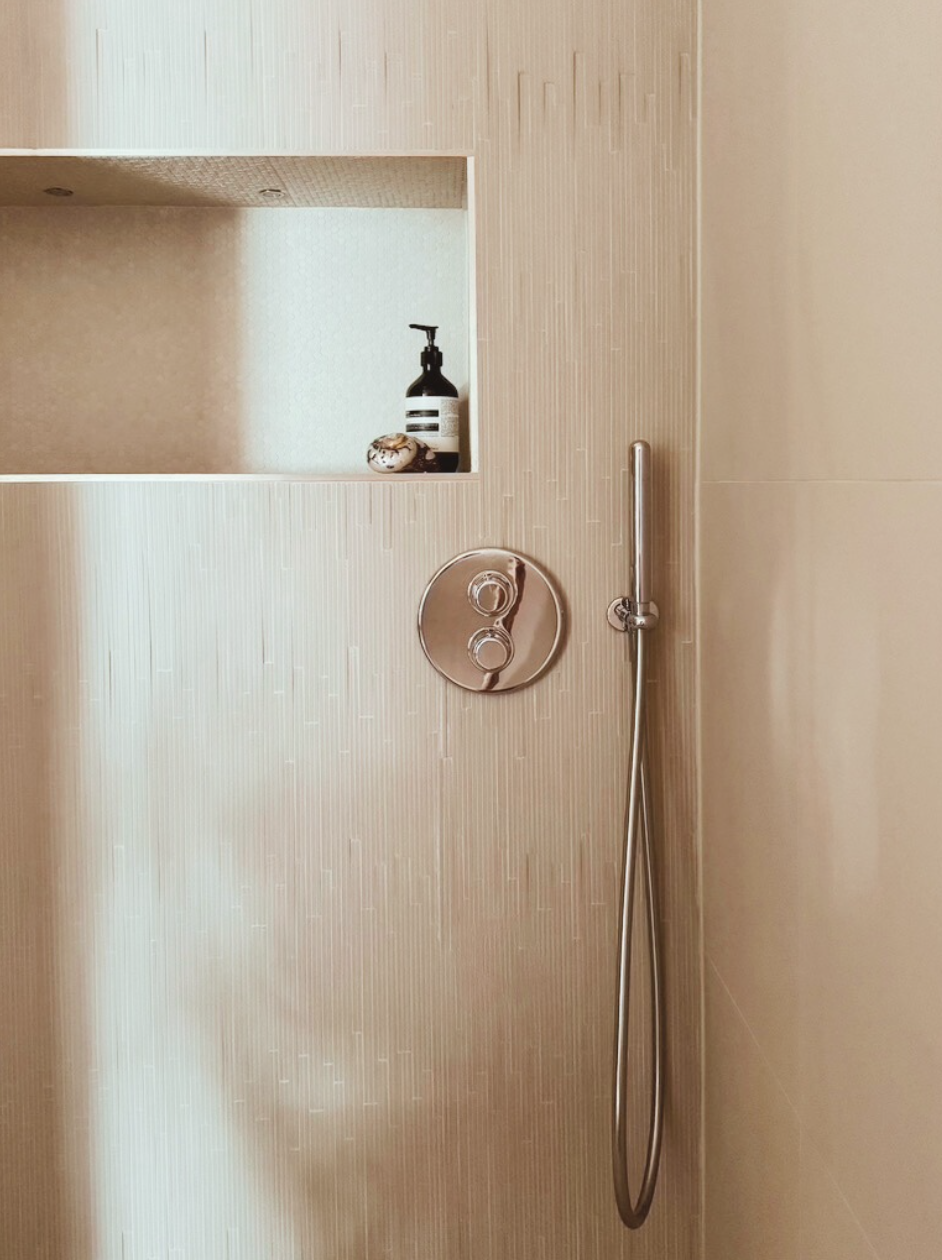 Close-up of a modern shower wall with a built-in shelf holding a soap dispenser and a small bowl, with a showerhead and control panel.