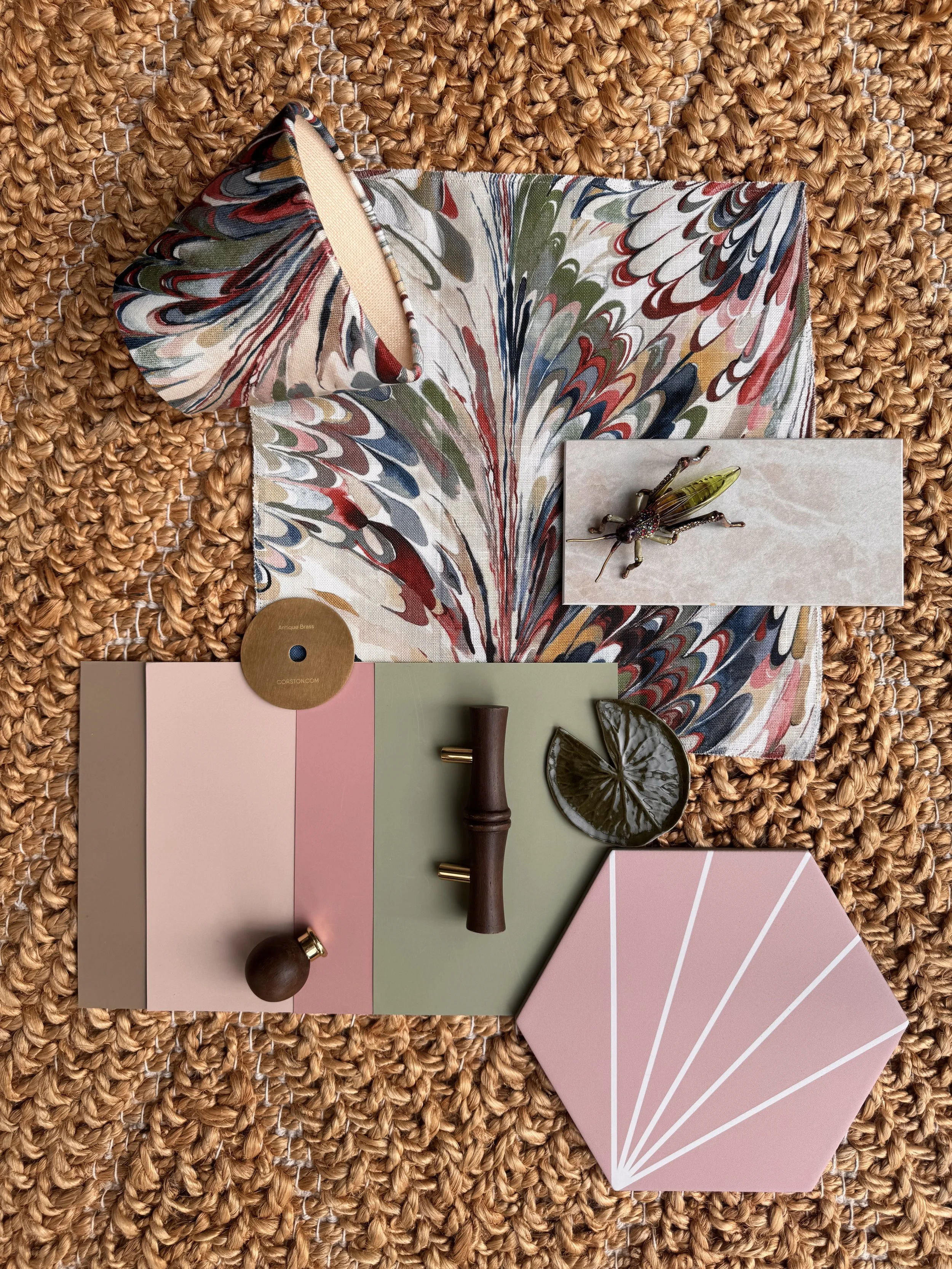 Various interior design materials including paint swatches in pink, beige, and green, a color fan deck, a small decorative insect figure, a dark wood and brass handle, a leaves-shaped coaster, and a strip of fabric with a colorful swirling pattern, arranged on a woven rattan surface.