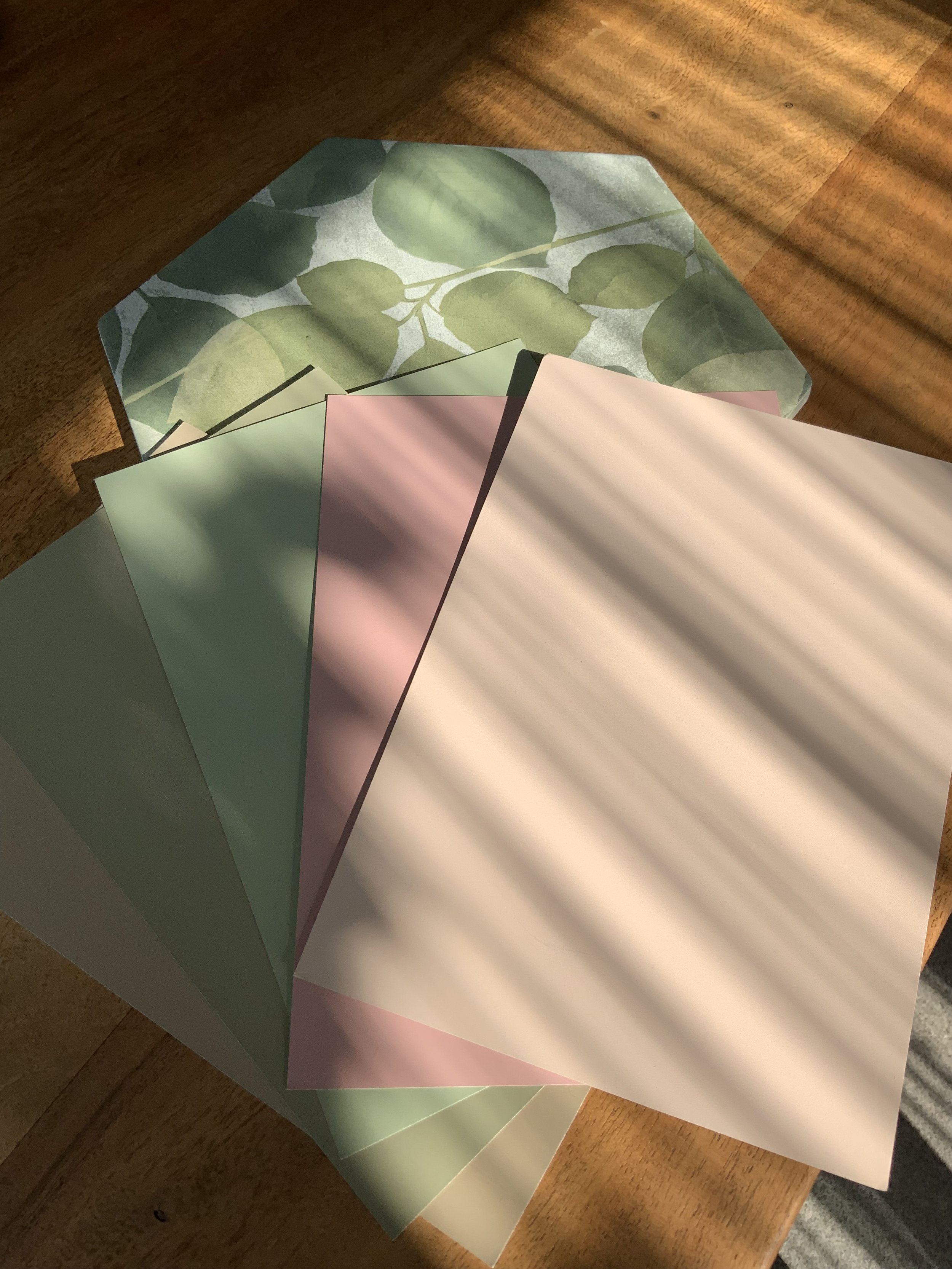Color swatches with pastel shades of pinks, greens, and beige laid out on a wooden surface, with a backdrop of leaf pattern paper.