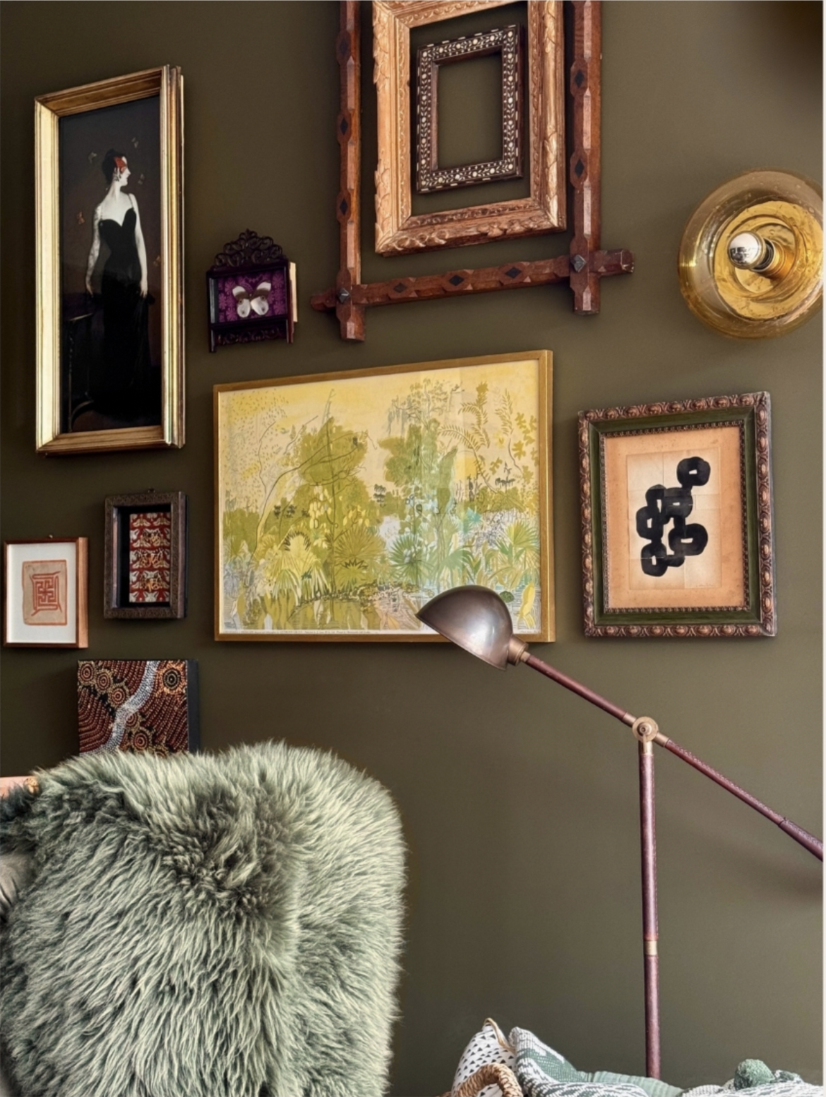 Olive green wall decorated with various framed artwork, including a portrait of a woman, botanical illustration, and abstract pieces, with a vintage-style floor lamp and a fuzzy green chair.