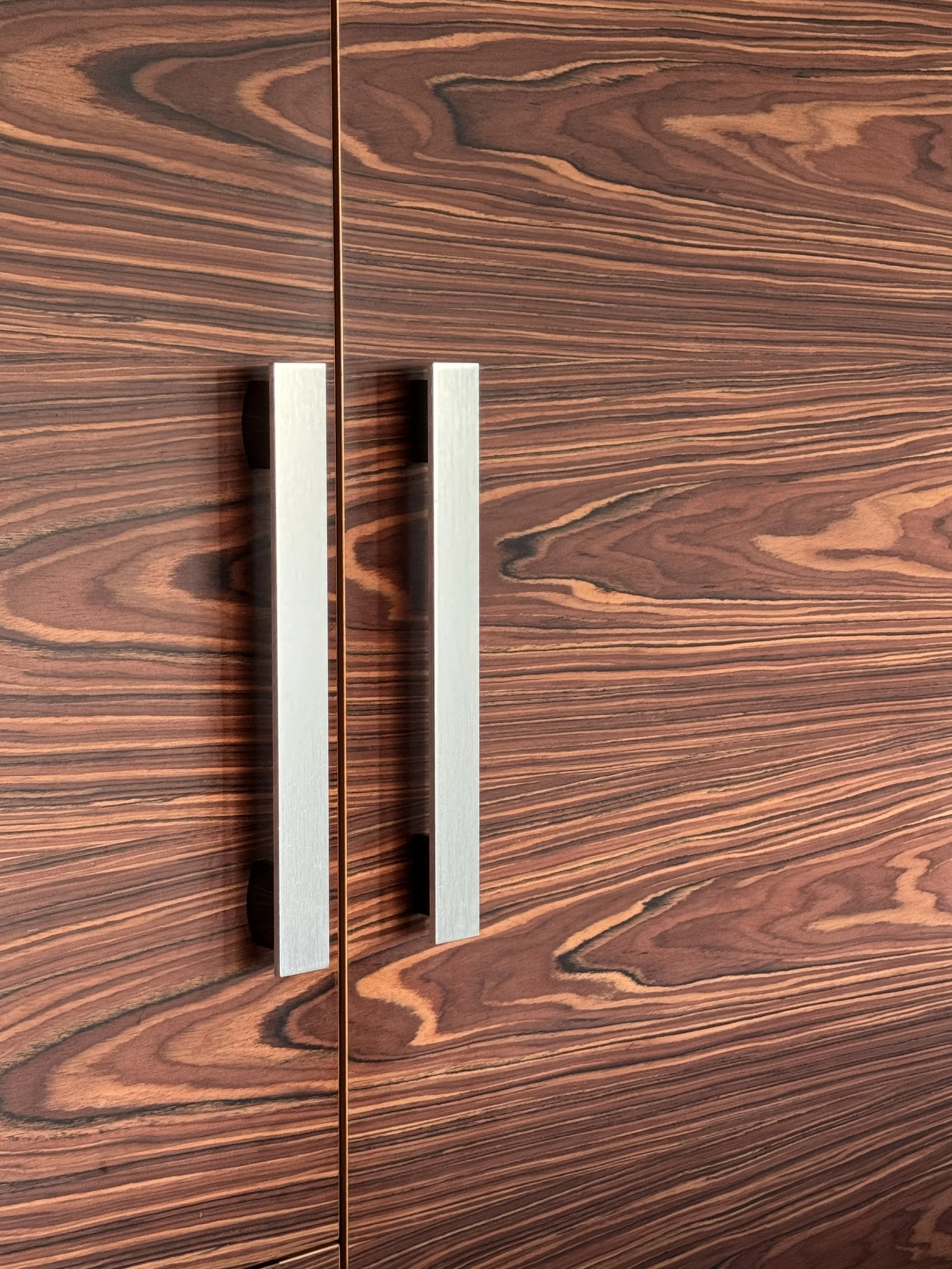 Close-up of a wooden kitchen cabinet with a dark wood grain pattern. The cabinet has vertical metallic handles.