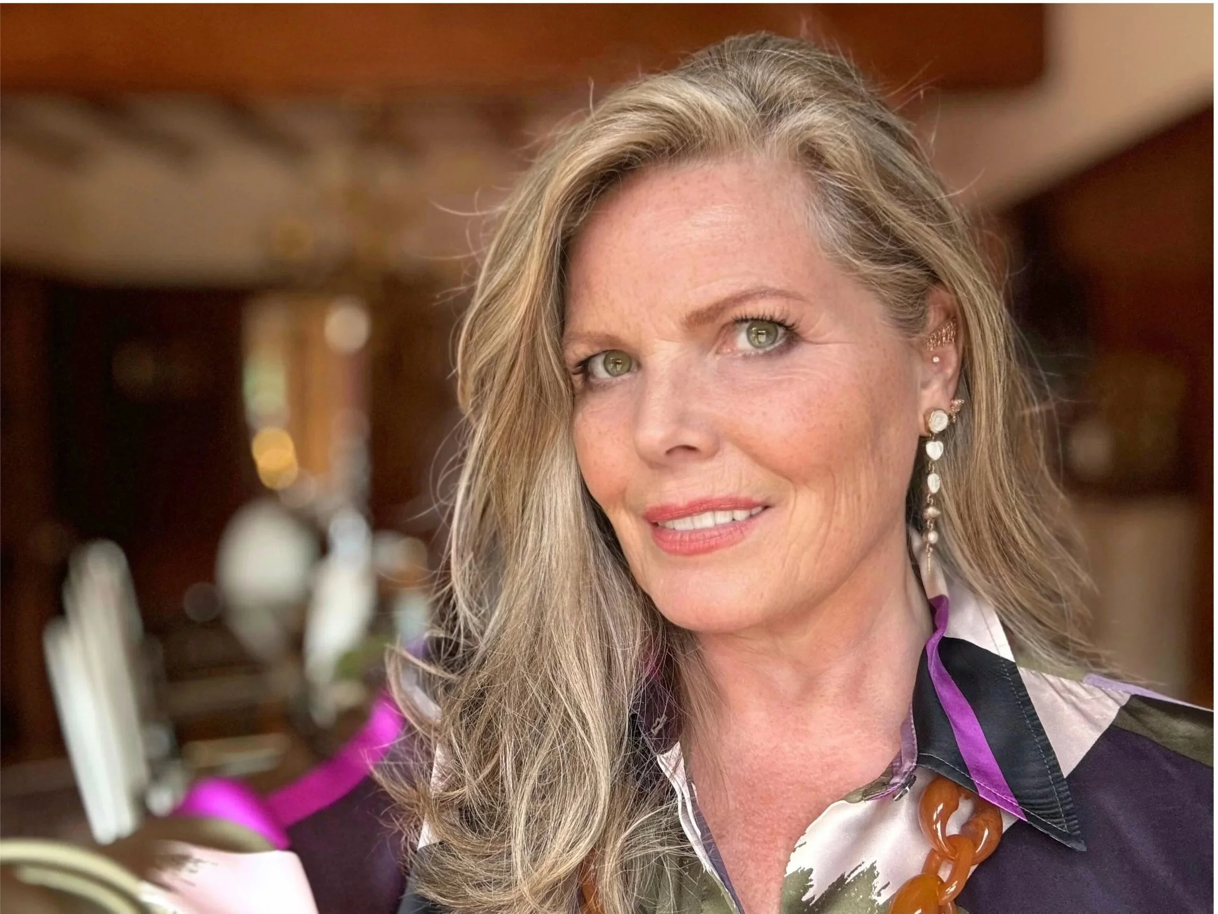 Close-up of a woman with long blonde hair, green eyes, makeup, earrings, and wearing a necklace and a black and purple striped garment, with a blurred background.