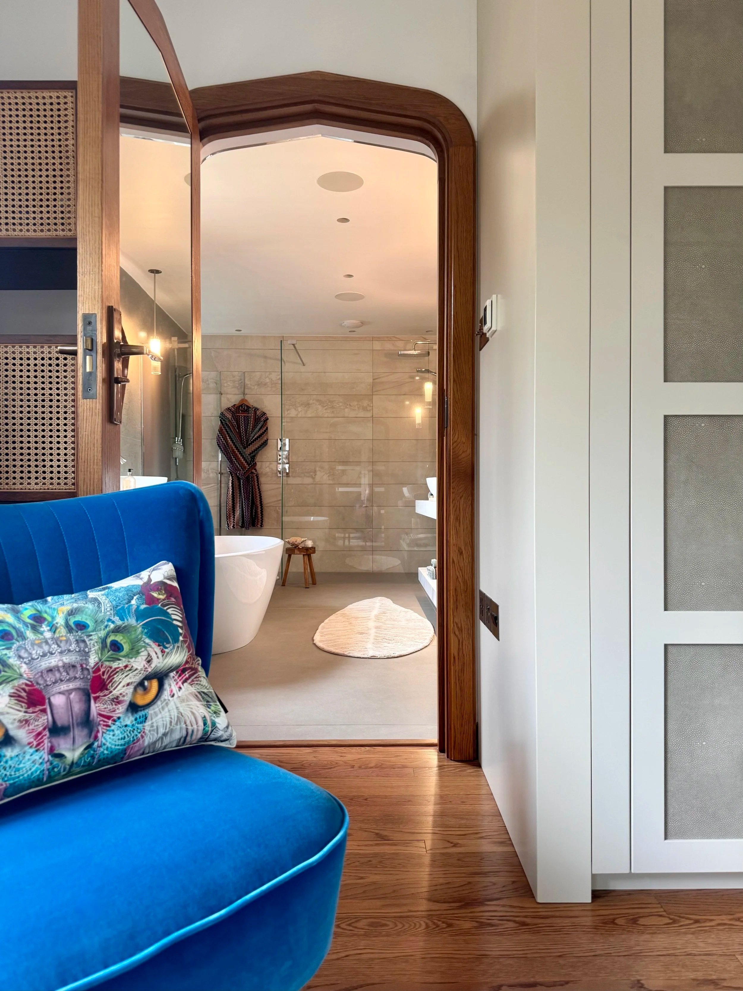 Interior view through an open wooden door into a room with wooden flooring, a blue velvet sofa with a colorful peacock feather pillow, a white bathtub, a small round beige rug, and a bathroom area with beige tiled walls and robes hanging.