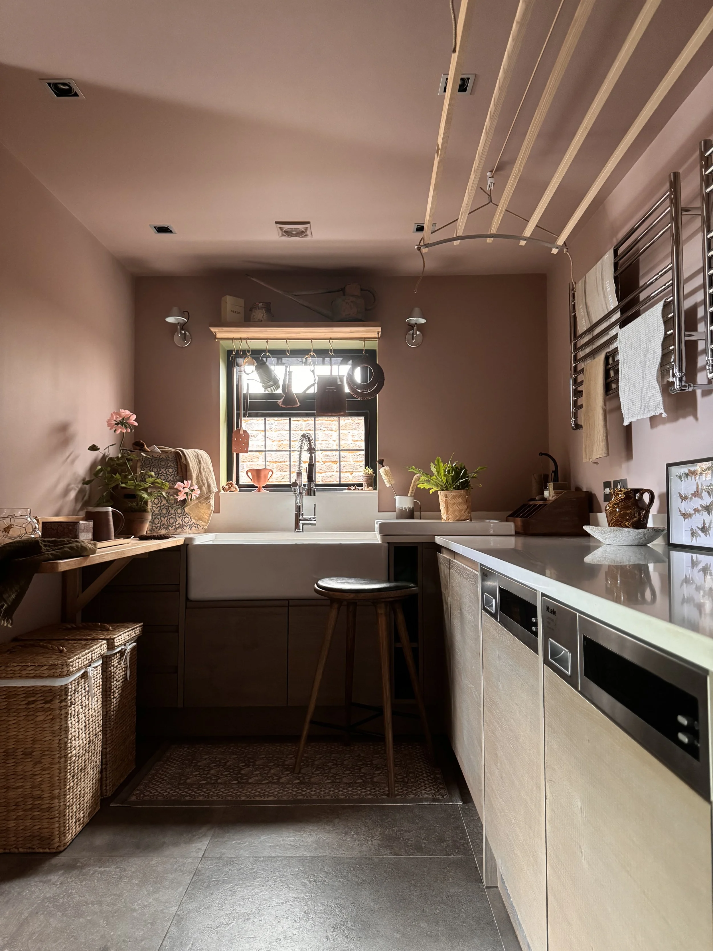 A cozy kitchen with a white sink under a small window, surrounded by beige cabinetry, plants, and kitchen utensils. Wicker baskets are on the left, with a wooden stool and a textured rug underneath. There's a dish towel hanging on a wall-mounted towel rack, and hanging from the ceiling are kitchen tools and wooden slats. The room has a modern rustic style with warm tones and practical decor.