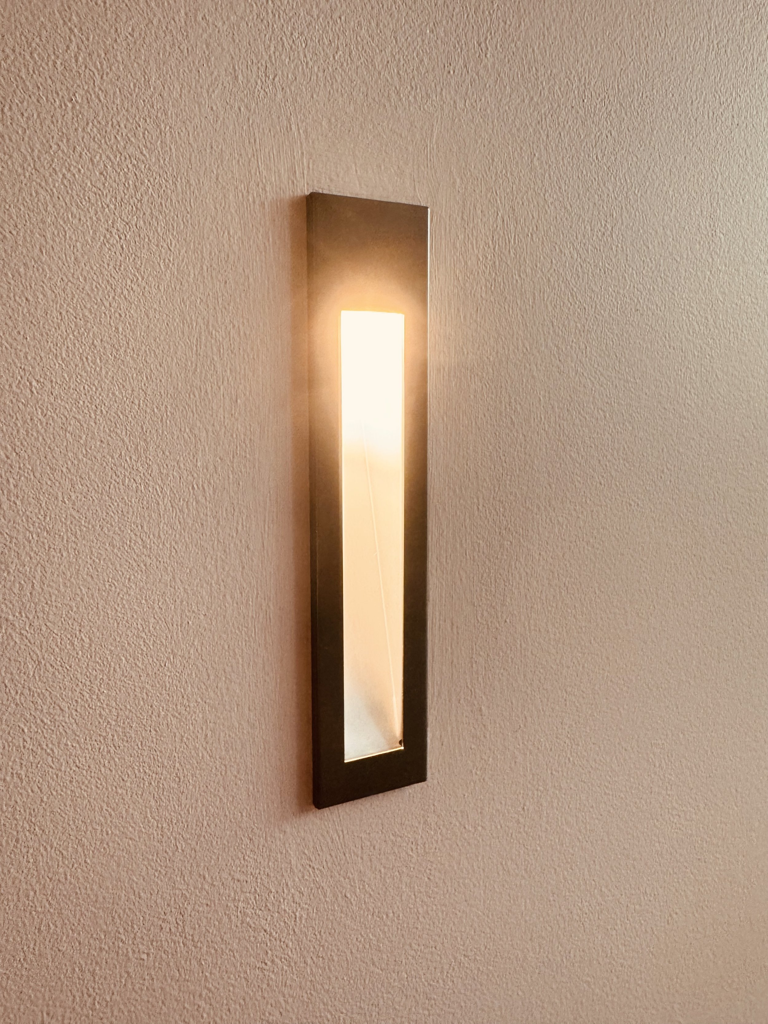 Wall-mounted modern light fixture with a rectangular design, emitting warm light against a textured beige wall.