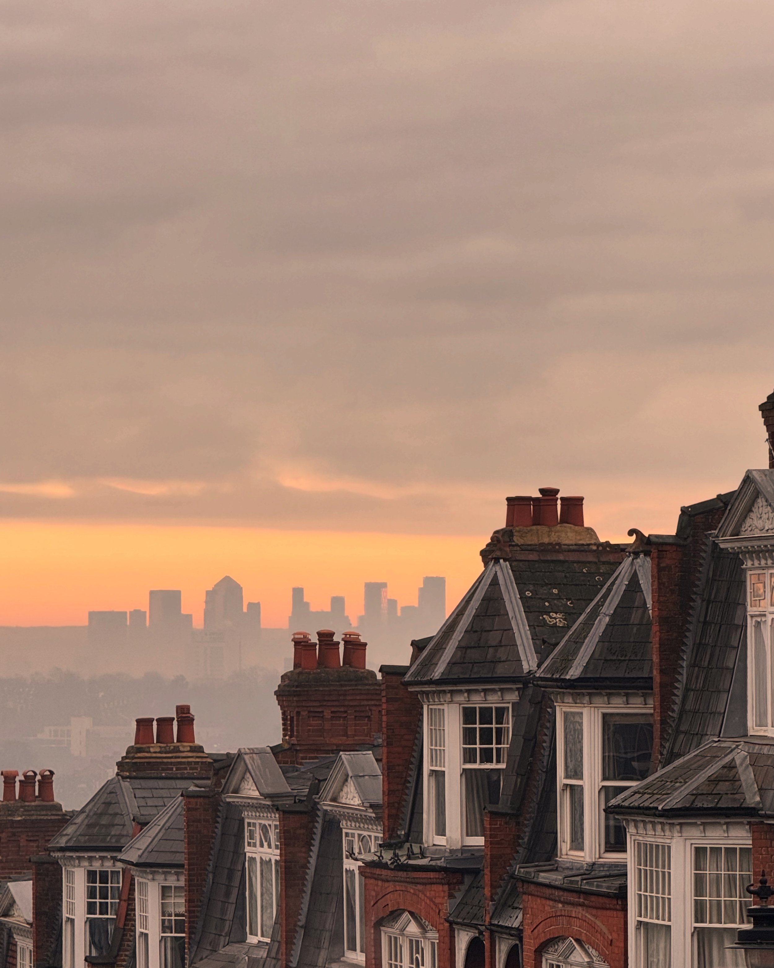 City skyline at sunset with the silhouettes of buildings and rooftops in Muswell Hill in the foreground, and taller skyscrapers of London City in the background under a cloudy sky.