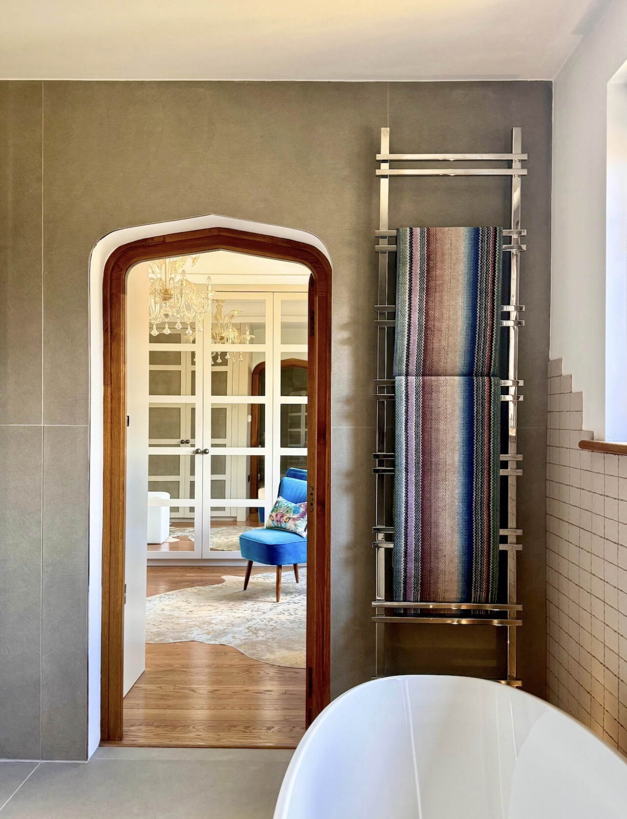View from a bathroom looking into a room through a rounded wooden doorframe. The room features a white cabinet, a blue armchair with a floral pillow, and a chandelier. The bathroom has beige and gray walls, a window, and a chrome towel rack with a striped towel.