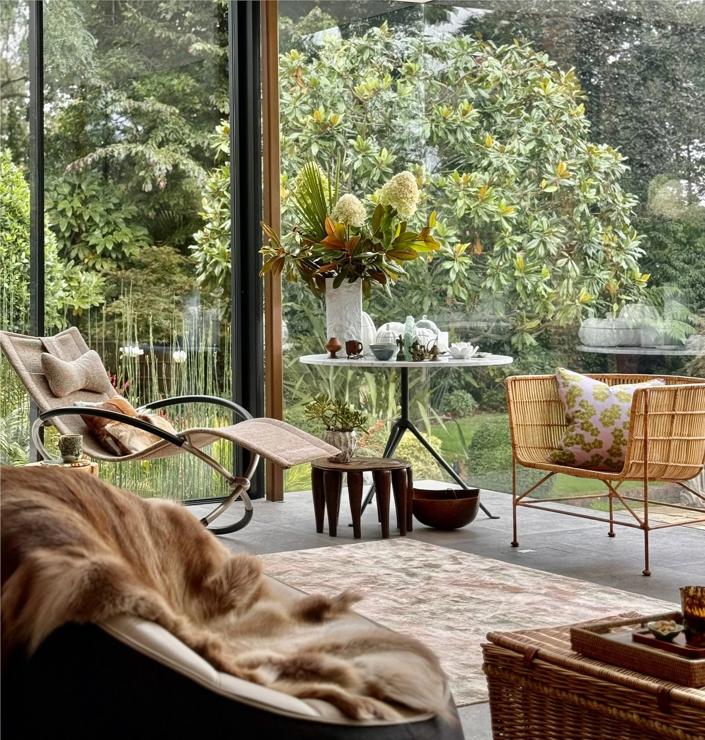 A cosy garden room seen from inside, a wicker chair with a patterned pillow, a round table with decorative items and a plant, and lush green trees and bushes outside through large glass