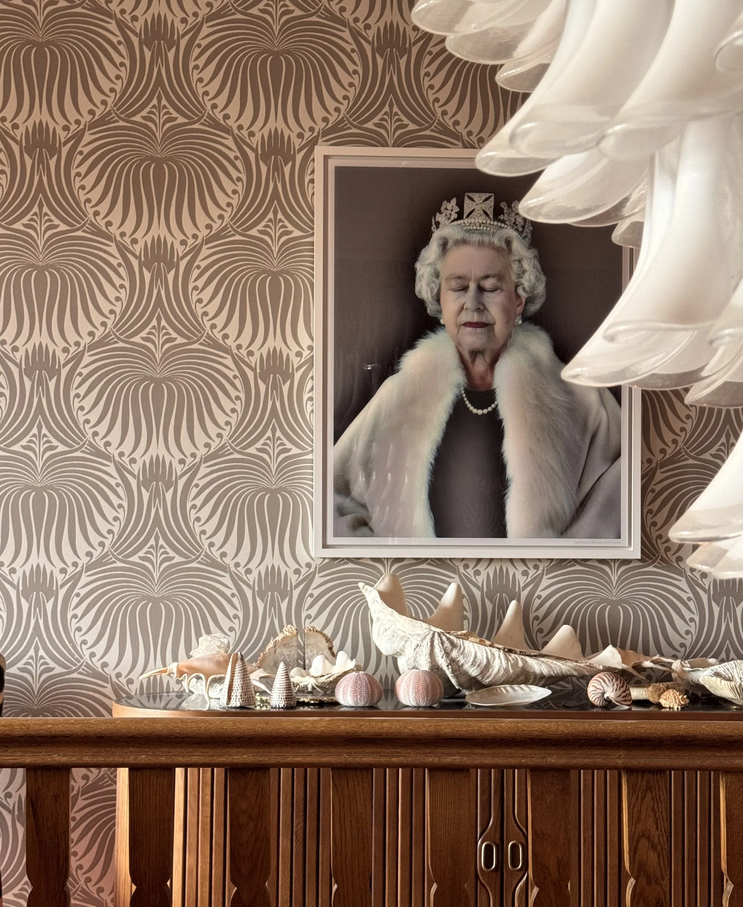 A framed picture of Queen Elizabeth II wearing a crown and a fur stole, with her eyes closed, hanging on a patterned wallpapered wall. In front of the picture, there is a wooden sideboard decorated with various seashells and coral.