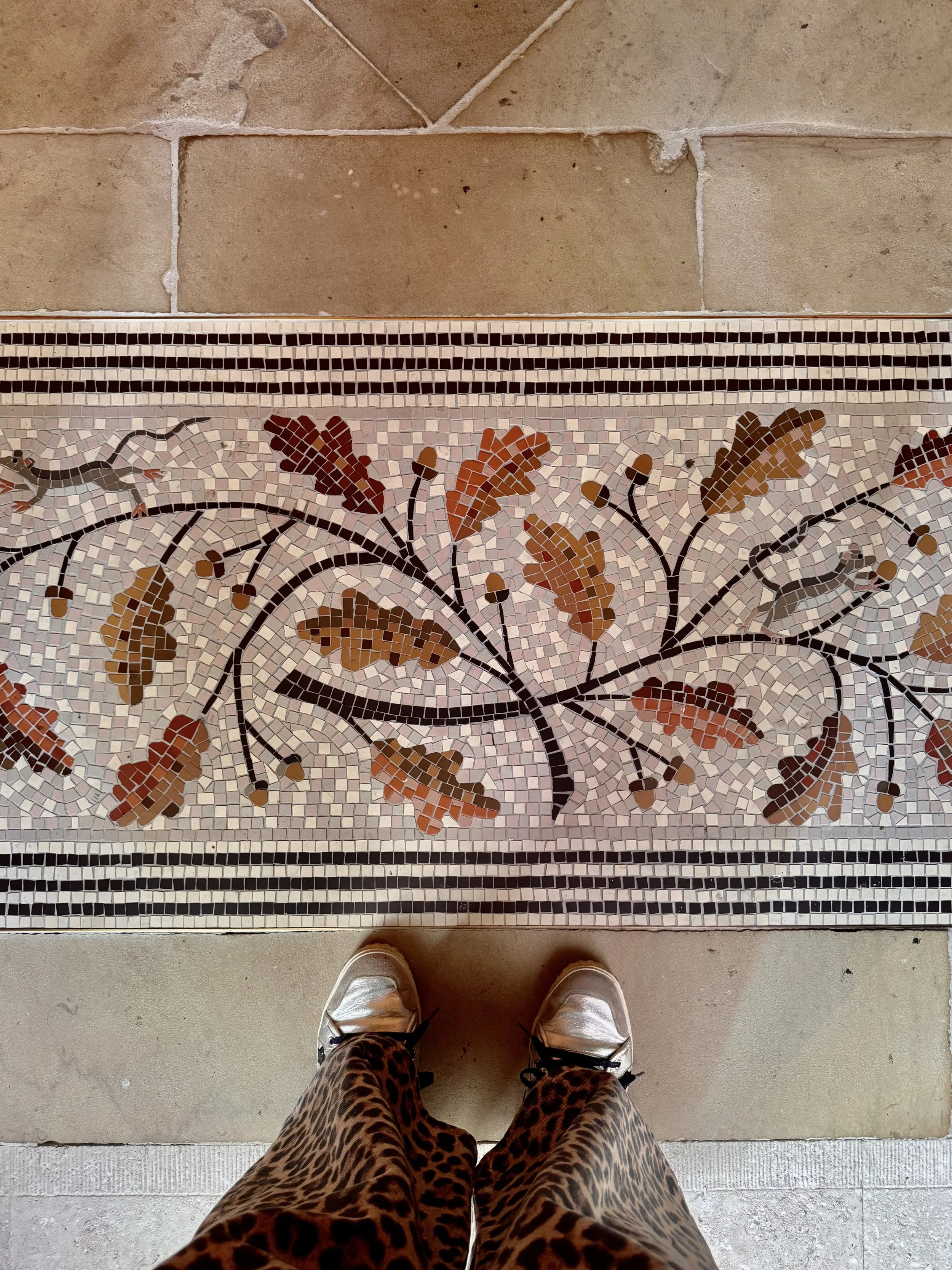 Photo from a top-down perspective of a person wearing leopard print pants and white sneakers standing on a concrete floor in front of a decorative mosaic tile design with floral patterns. The person's feet are visible, and they are wearing white shoes with black laces.