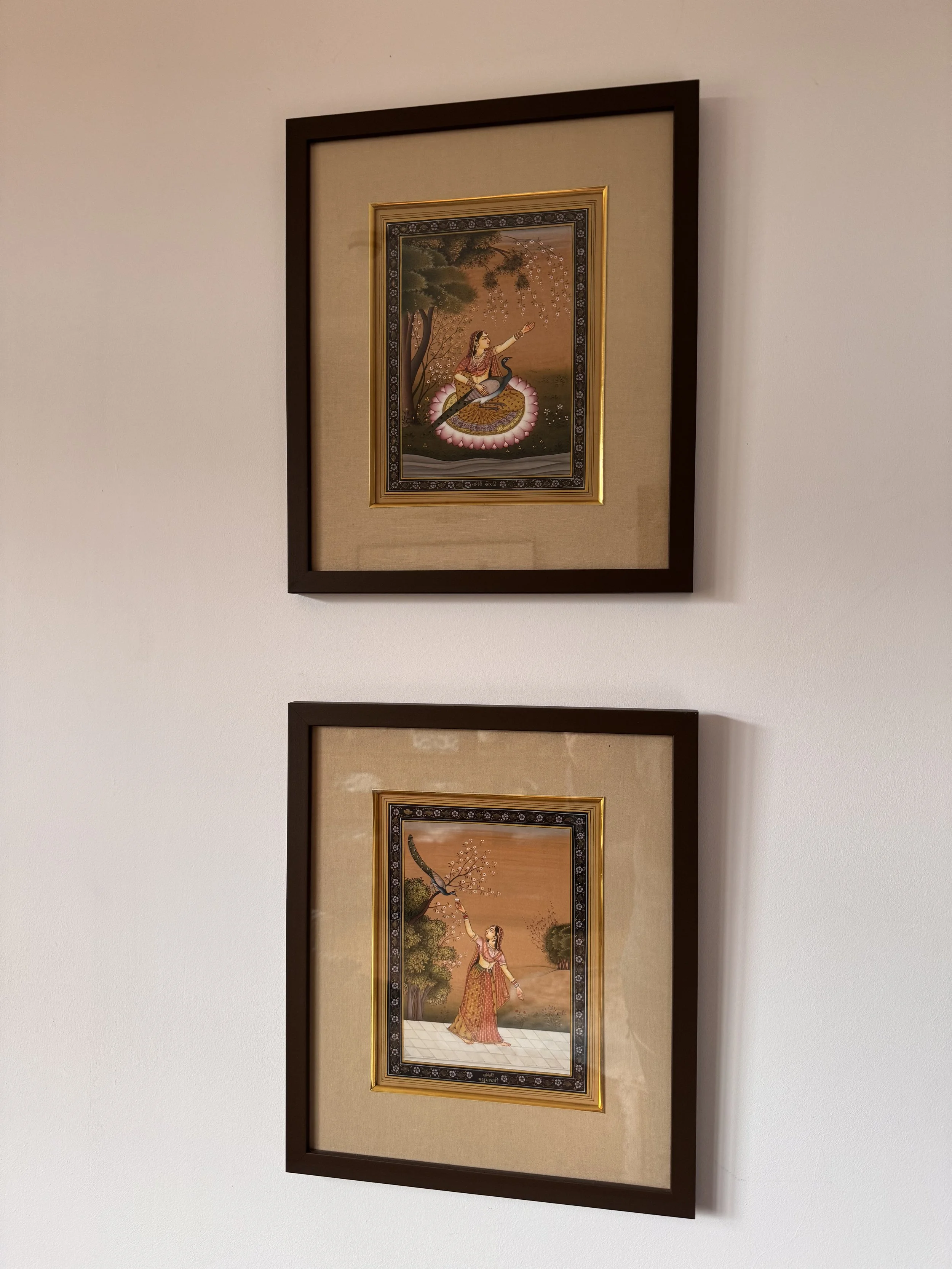 Two framed traditional Indian paintings with gold accents on the frames, hanging on a white wall. Each painting depicts a woman performing classical dance in natural settings.