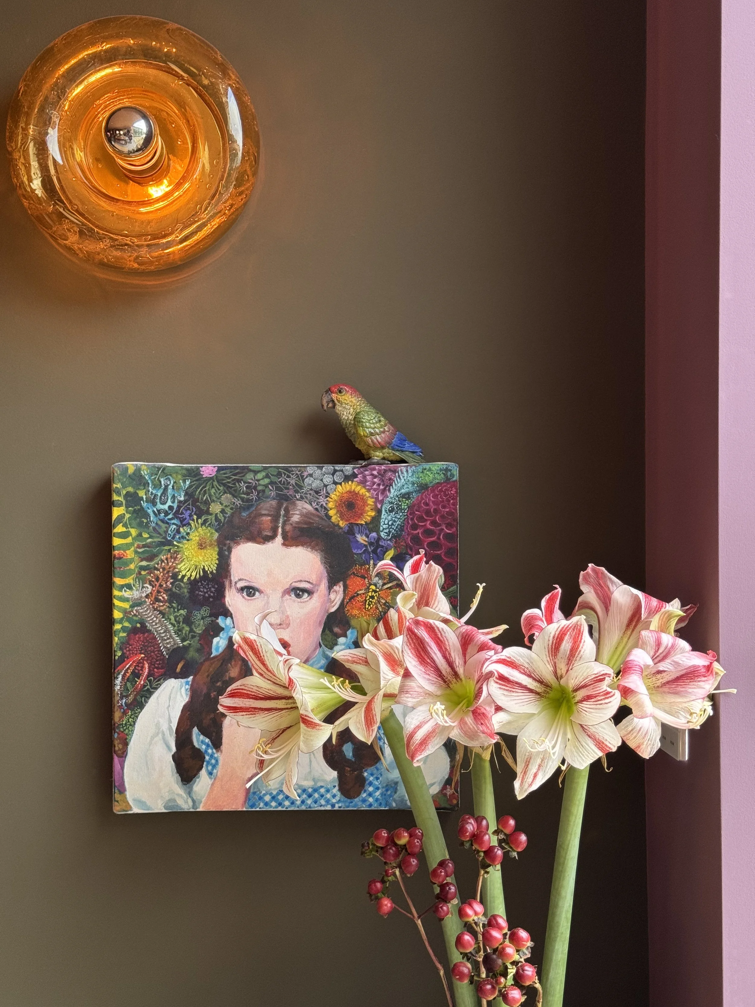A colorful framed portrait of a woman with dark hair and a white shirt, surrounded by vibrant flowers and a green plant, with a brown wall and hanging decorative light in the background.
