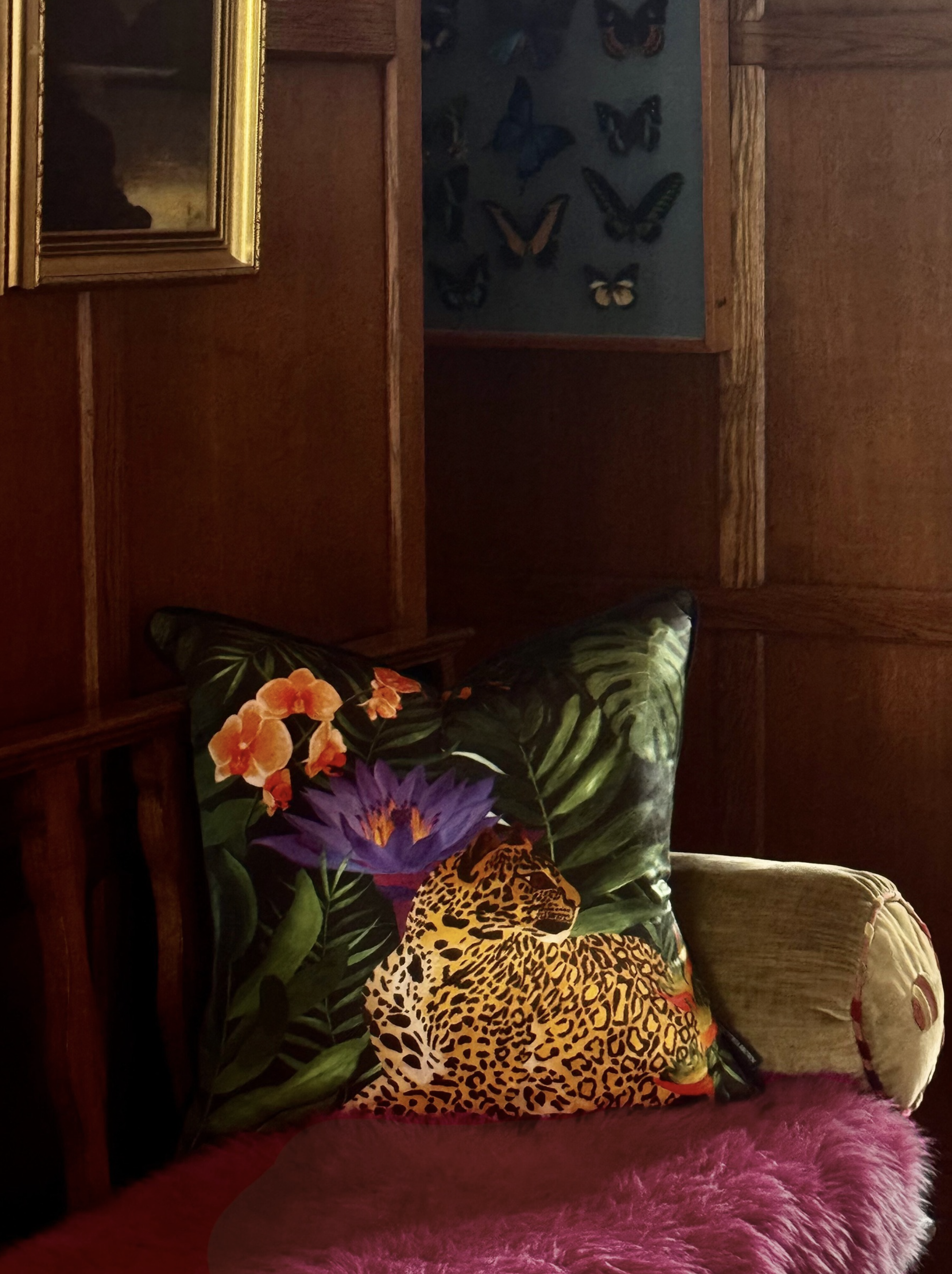 A corner of a room with a wooden frame, the bench has vibrant animal-print and tropical-themed pillows. A pink sheep skin throw is on the seat, and a floral cushion with large purple and orange flowers and green leaves is partially visible.