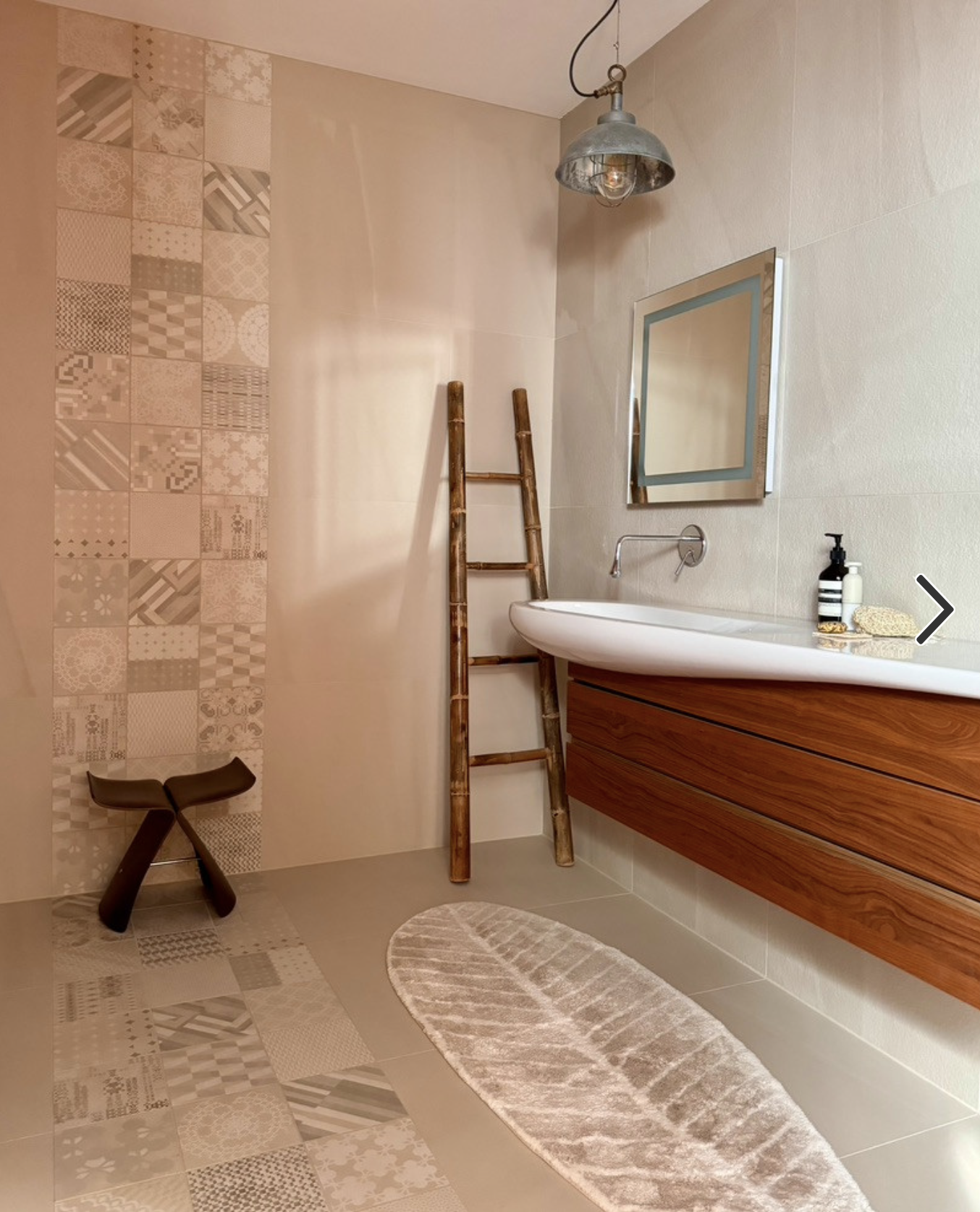 A modern bathroom with a wall-mounted mirror, a round vessel sink with a wooden vanity, a bamboo ladder, a metallic pendant ceiling light, a patterned towelling rug on the floor, and decorative bottles and a sponge on the sink counter.