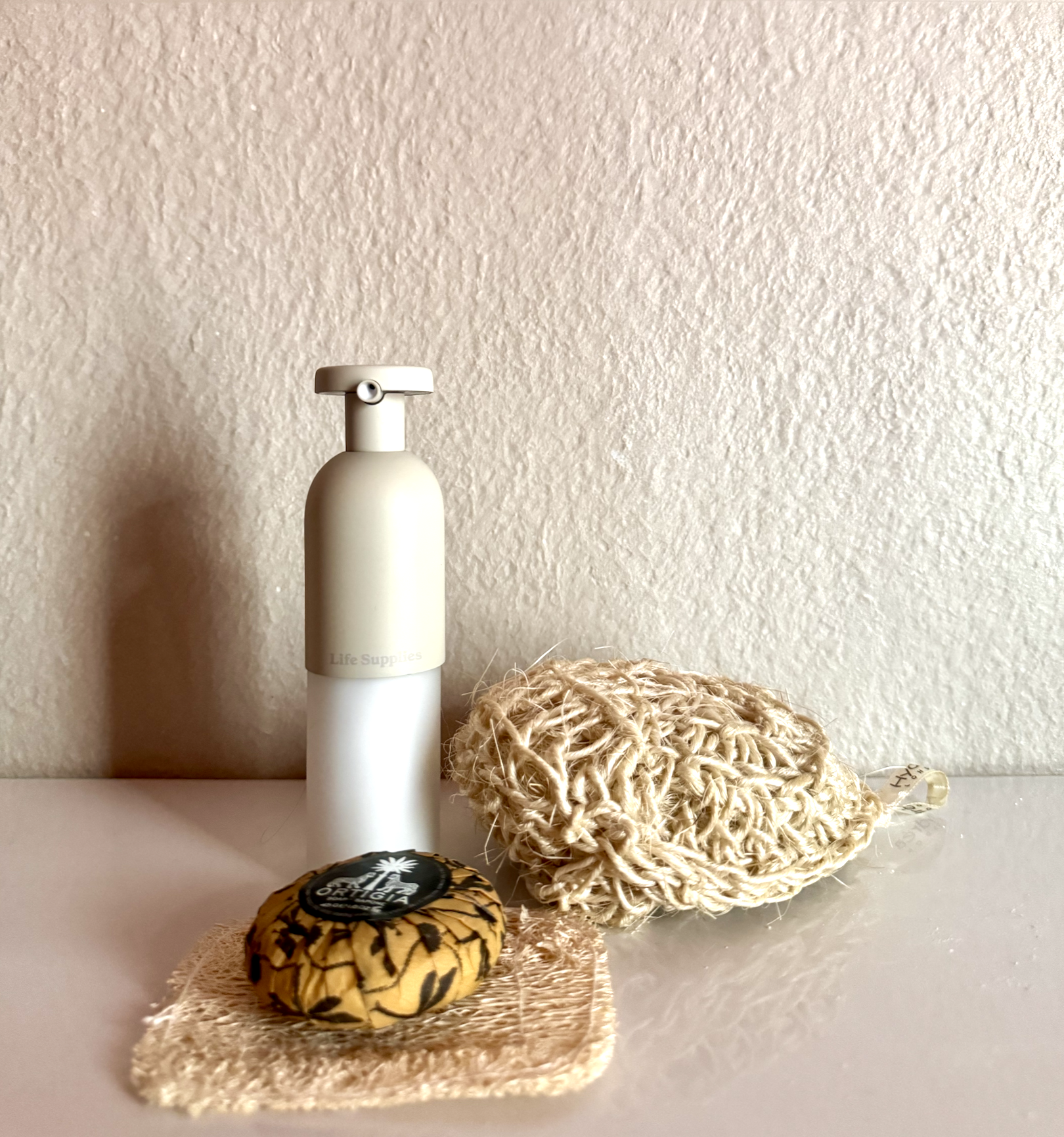 A white bottle of skincare or cosmetic product, a round decorative object with a black label, a beige woven scrubby pad, a pink textured cloth, and a small white container, all arranged on a flat surface against a beige textured wall.