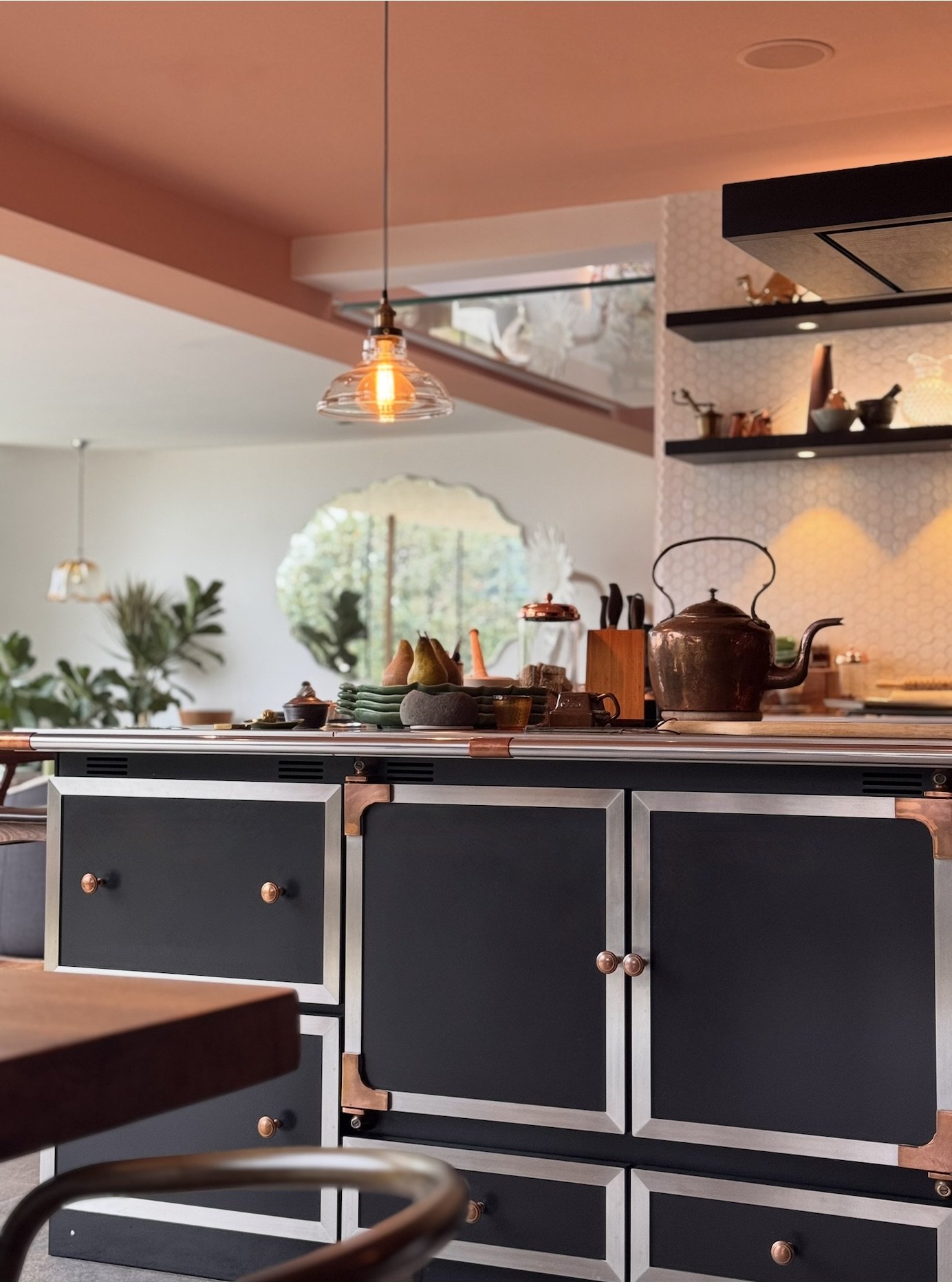 Kitchen with La Cornue island, copper accents, a countertop with decorative items, including a teapot and fruit, under warm lighting. Open shelving displays bowls and small decorative objects, with a large window in the background showing greenery.