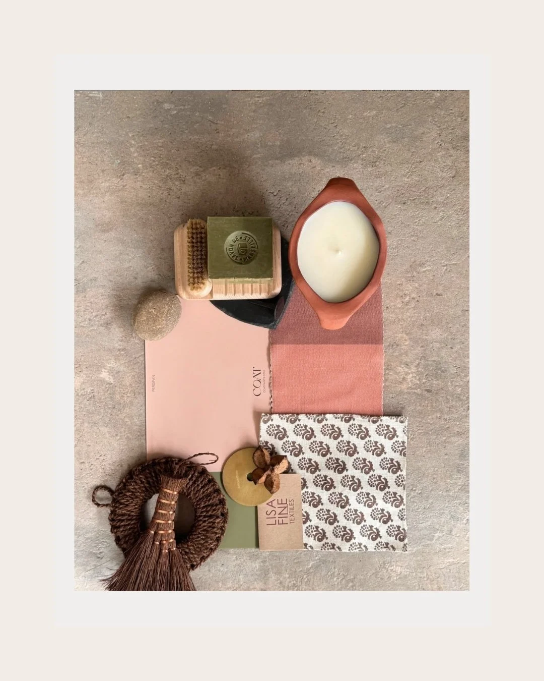 Flat lay of various natural and minimalist home decor items including a candle, soap, textured ball, wooden brush, cloth with floral pattern, and pink and beige fabric swatches, arranged on a pink sheet.