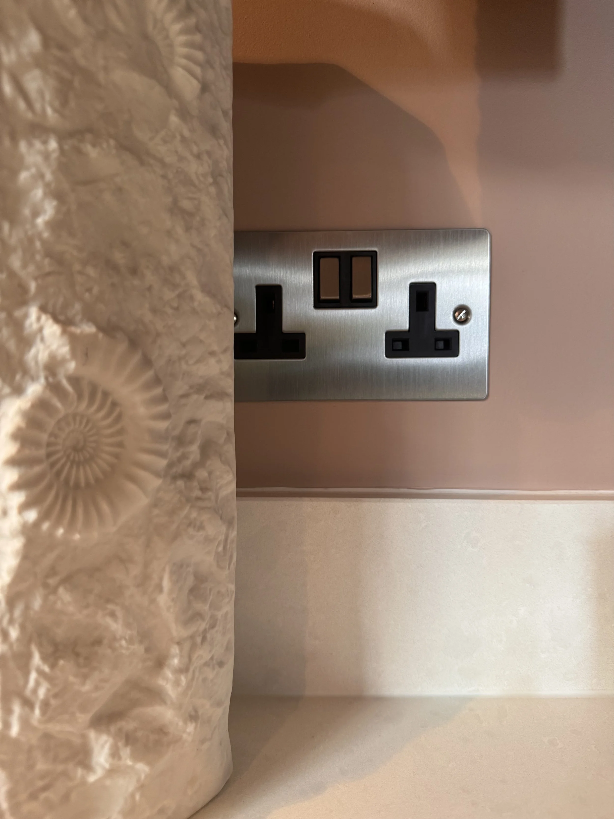 Close-up of a beige textured vase with spiral shell patterns and a corner of a room with pink walls and a metal electrical outlet with two black sockets.