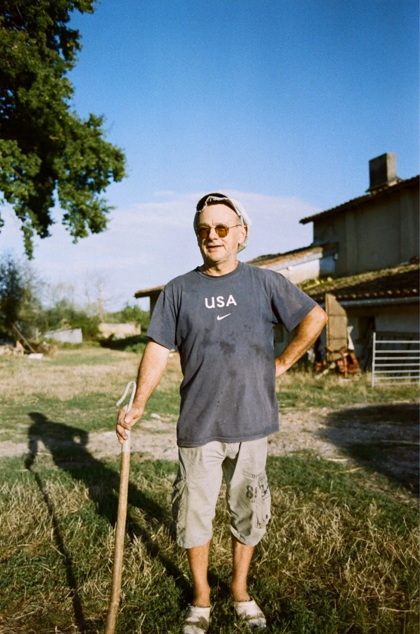 Homme debout dans un jardin, portant un t-shirt gris avec l'inscription 'USA' et une Nike, des lunettes de soleil, un chapeau blanc, tenant une pelle, avec une maison en arrière-plan sous un ciel bleu clair.
