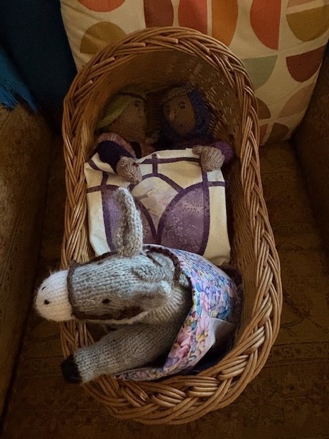 Wicker doll cradle with handmade stuffed animals inside, including a rabbit and a dog, with cushions in the background.