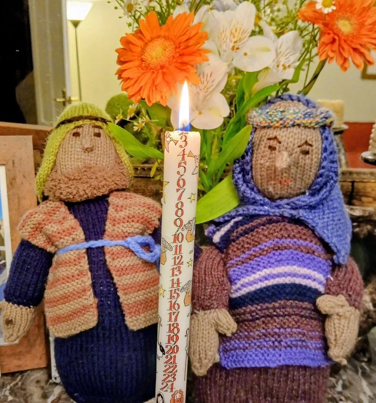 Knitted dolls of Mary and Joseph, a lit birthday candle, and a bouquet of flowers on a table.