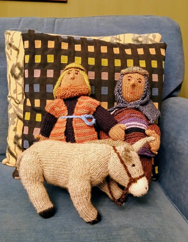 Knitted dolls of a man and a woman riding a knitted horse on a blue couch, with decorative pillows in the background.