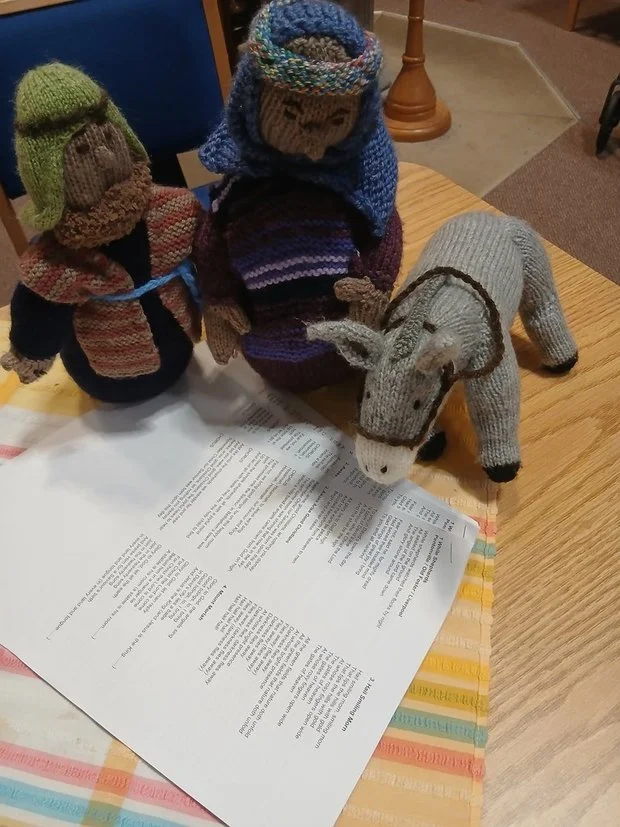 Two handmade knitted dolls dressed in winter clothes, a stuffed animal horse, and a sheet of paper on a wooden table.