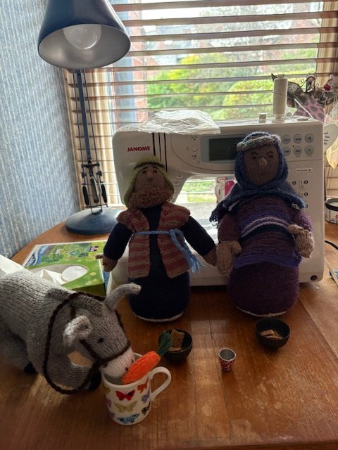Handmade dolls, a stuffed animal, and sewing supplies on a wooden table in front of a window with blinds and a sewing machine.