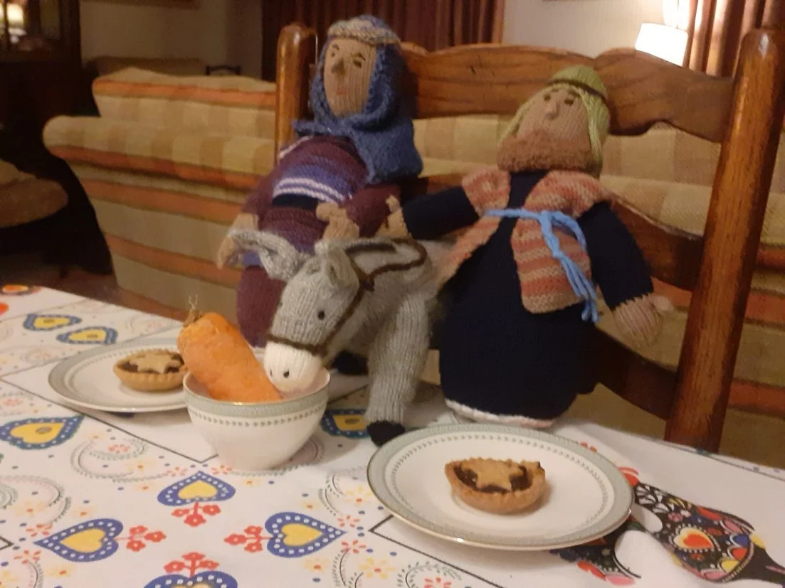Two sock dolls dressed in winter clothing sit on a table with plates of cookies and a bowl with a large carrot.
