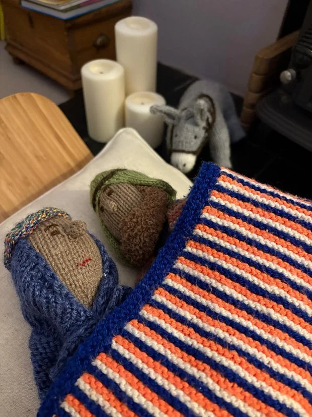 Three knitted stuffed animals resembling people are on a chair, with a striped blanket partially covering them. In the background, three white candles and a small stuffed donkey are on a table.
