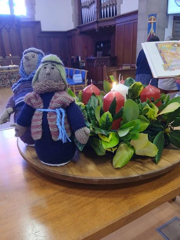 Knitted nativity figurines of Mary and Joseph on a wooden table with Advent candles and greenery, inside a church.
