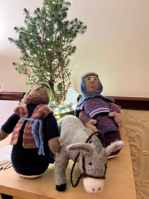 Handmade knitted dolls and a stuffed donkey figurine sit on a wooden table, with a small Christmas tree decorated with lights in the background.