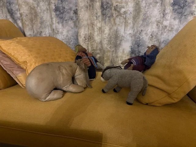 A bed with yellow pillows and mustard-colored bedsheets arranged with stuffed animal toys, including a dog, a bear, and dolls, on top.