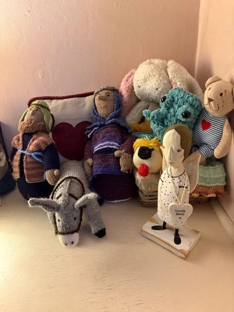 Collection of stuffed animals and handmade dolls on a white surface against a beige wall, including a gray and white donkey, a pink and white bunny, a blue plush toy, and a small figurine of a girl with puppy ears.